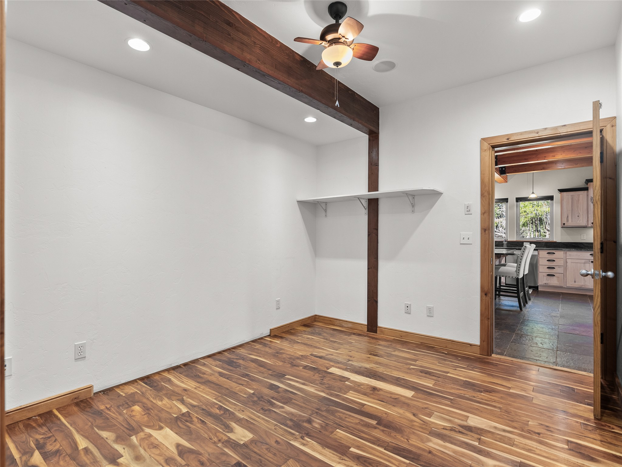 Unfurnished room with patterned wood flooring, exposed beam, ceiling fan, and doorway to the kitchen.