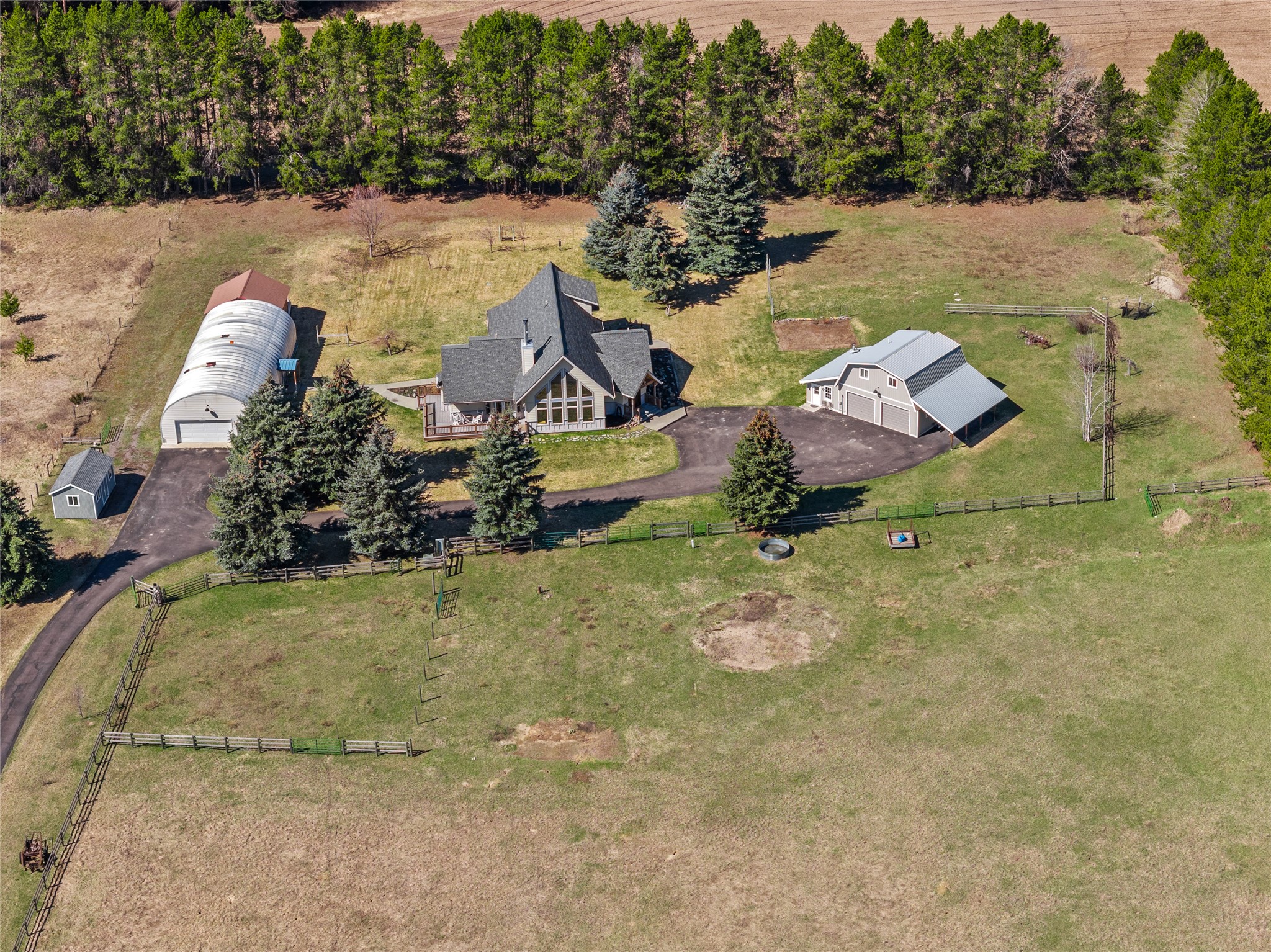 Aerial view of a rural home with large outbuildings, fenced pasture, and evergreen trees.