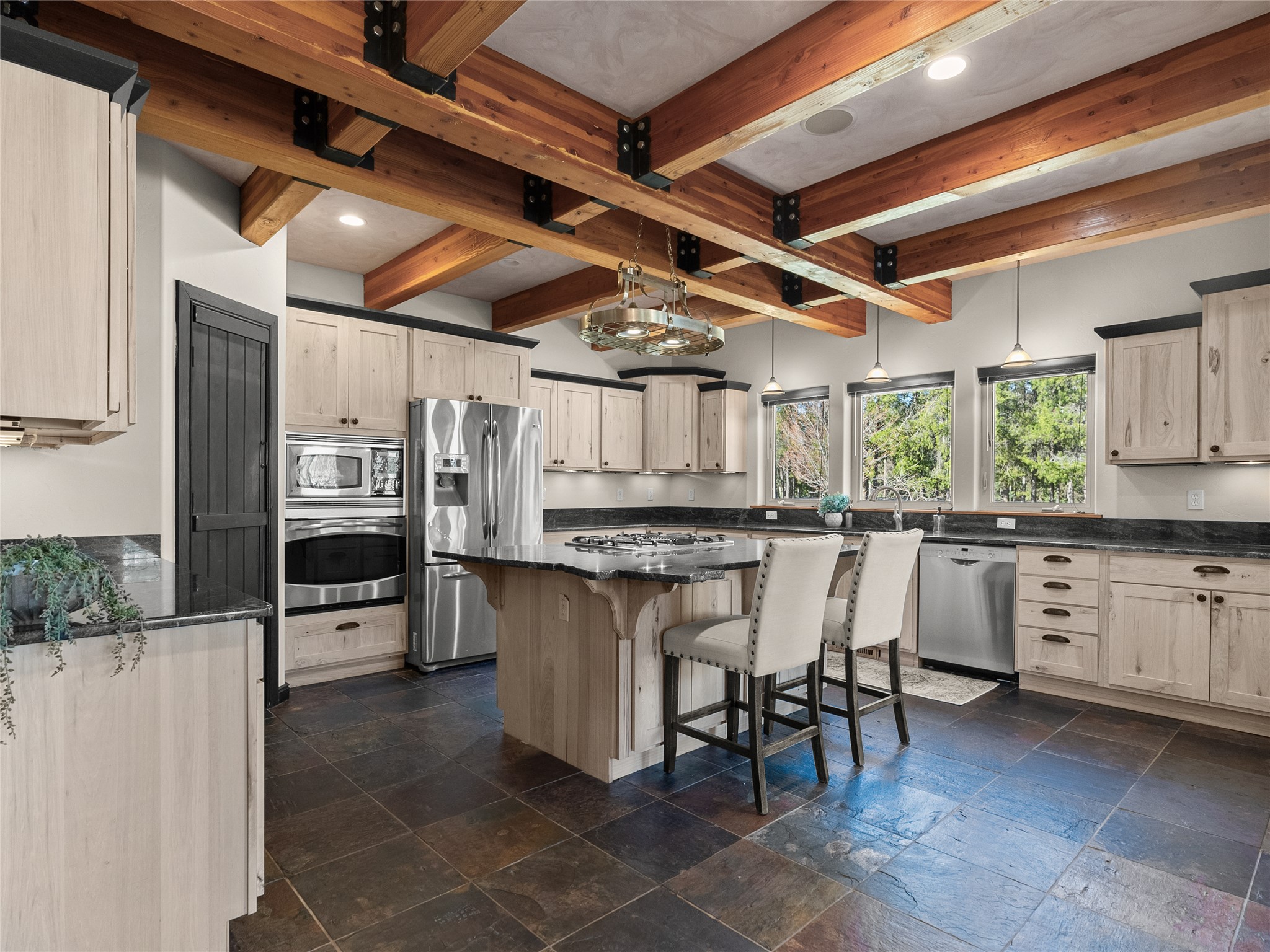 Kitchen with exposed timber beams, a stone island, stainless steel appliances, and windows overlooking trees.
