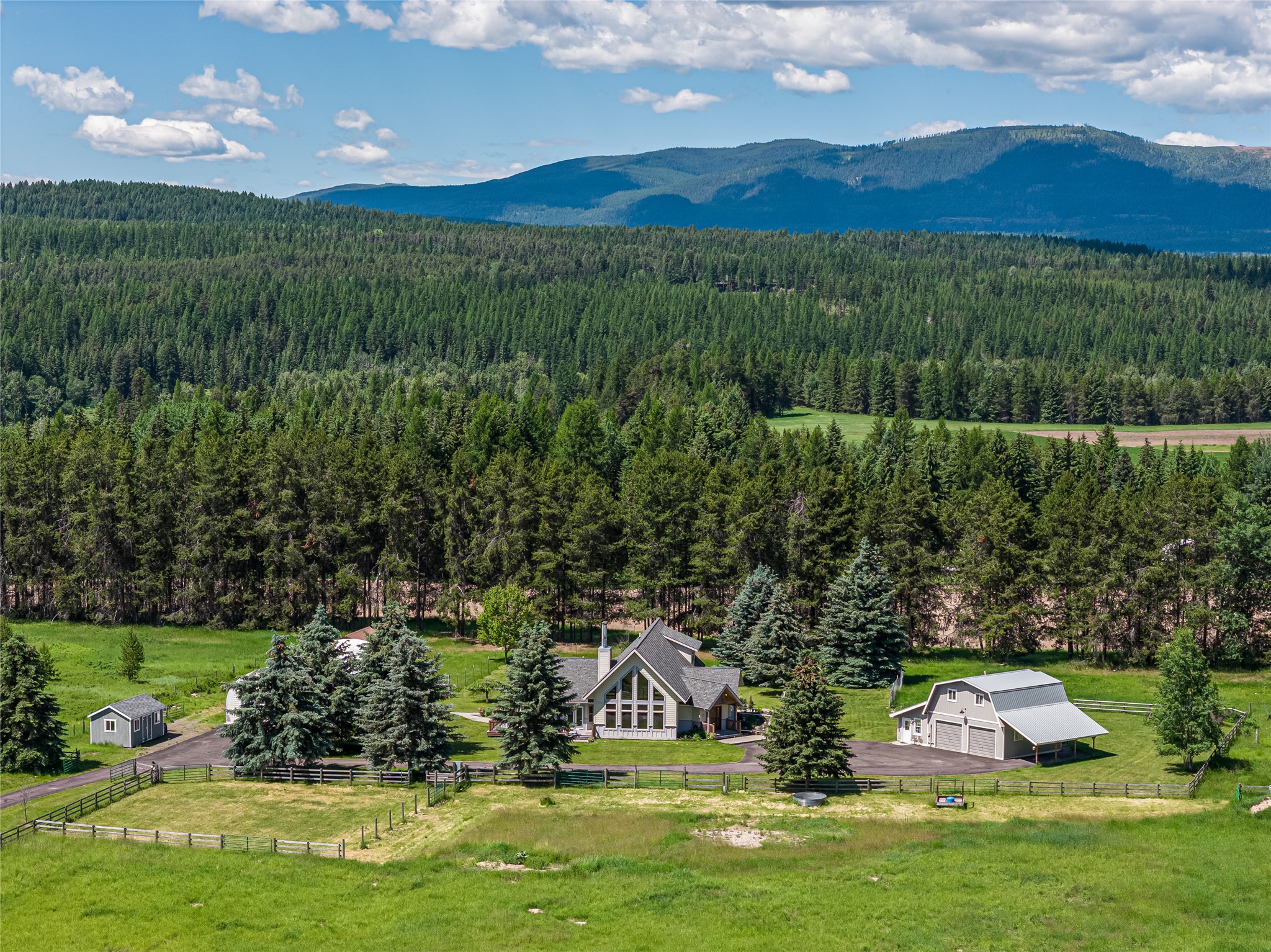 Rural home and outbuildings amid fenced green grounds, evergreen forest, and distant mountain ridges.