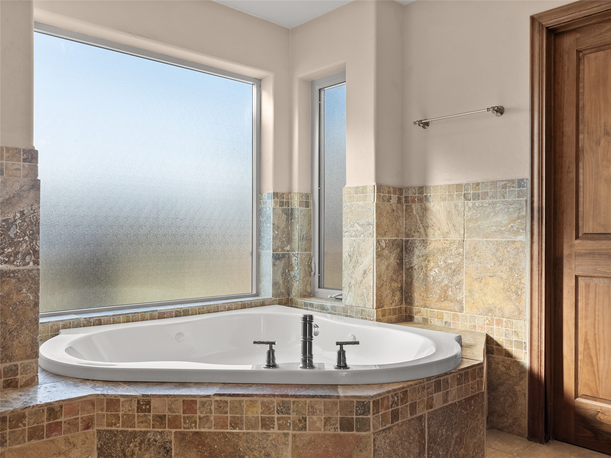 Corner soaking tub with stone tile surround beneath two frosted windows.