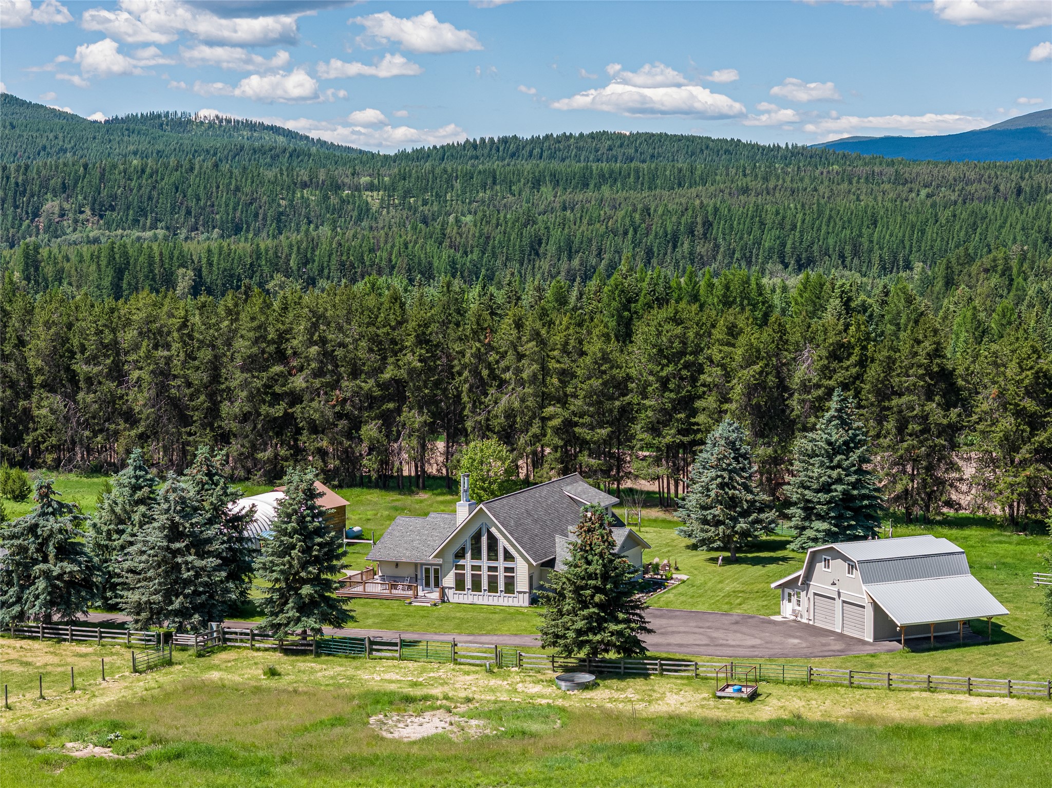 Aerial view of a home and detached outbuilding amid lawns, evergreen forest, and mountain ridges.