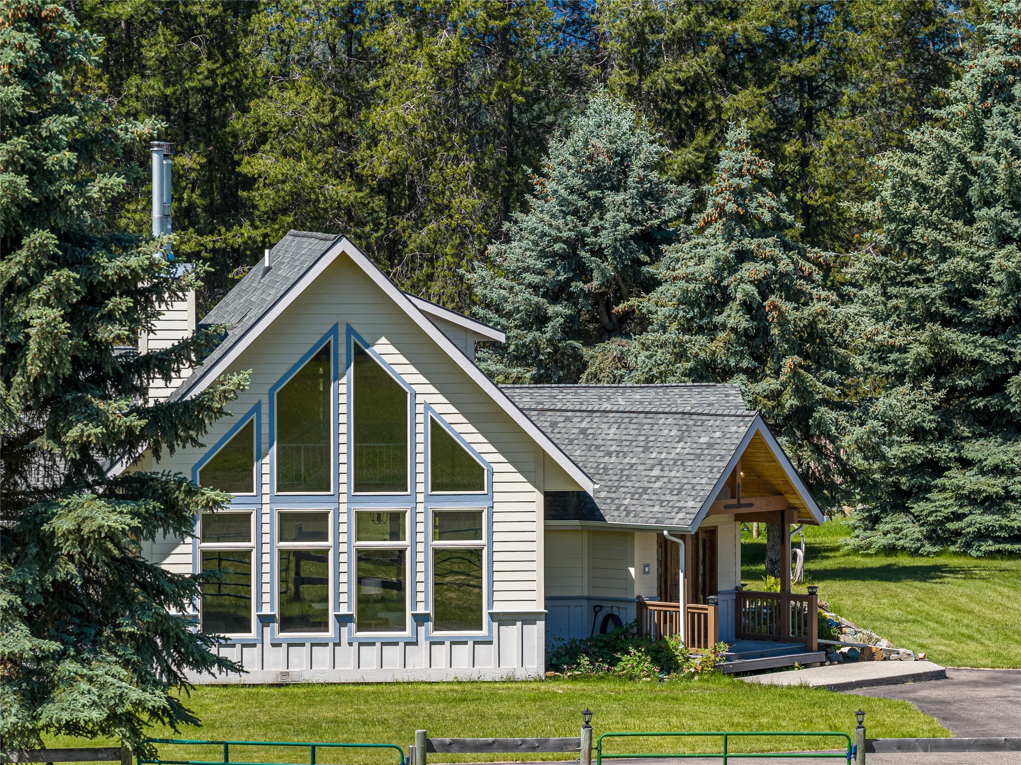 Light-colored house with tall gabled windows and a covered wooden porch among evergreen trees.