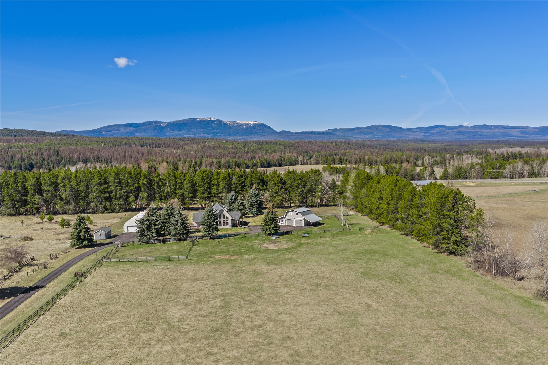Aerial view of a rural home and fenced acreage, with evergreen forest and distant mountains.