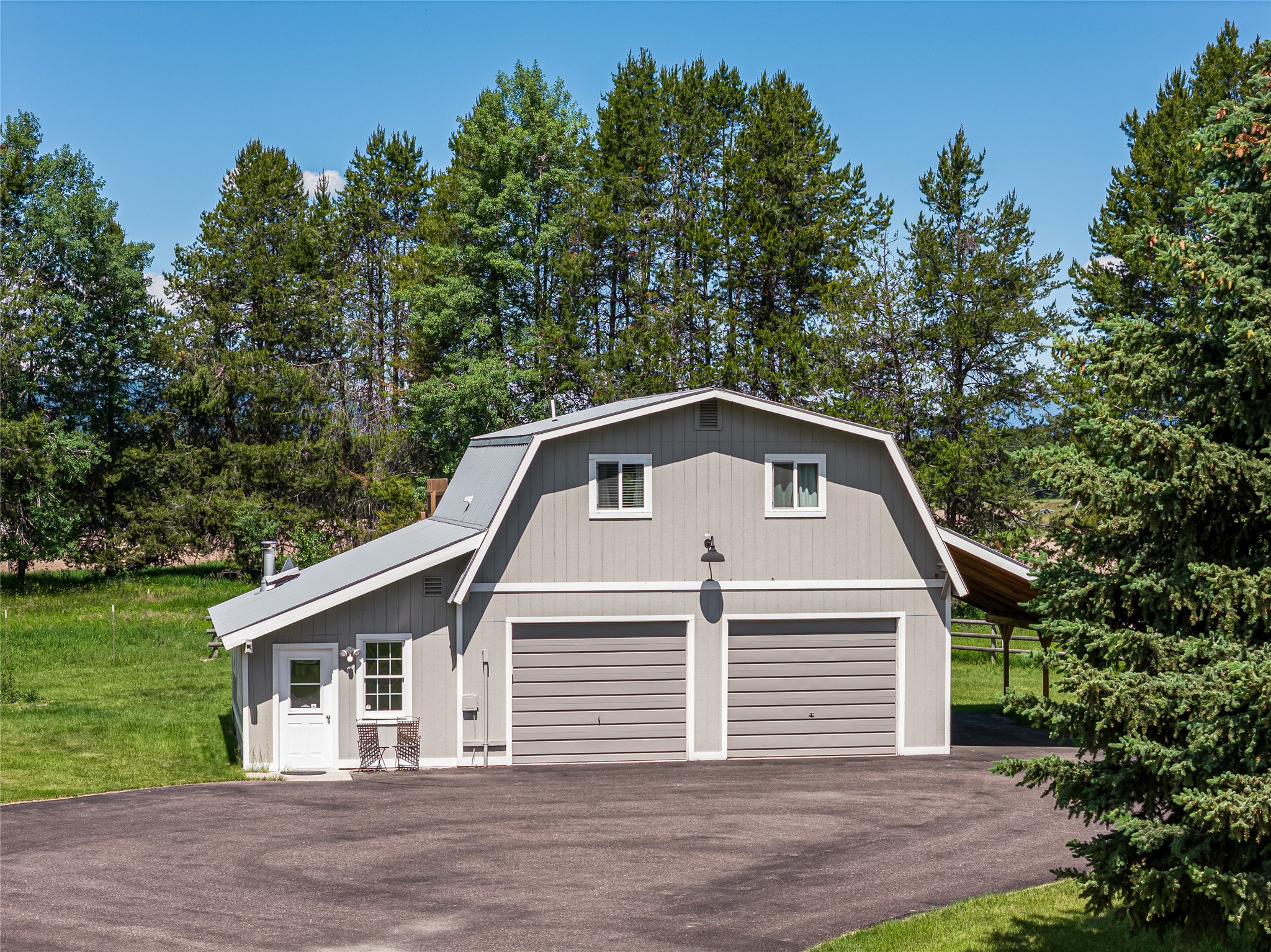 Gray barn-style outbuilding with two garage doors and a paved driveway among evergreen trees.