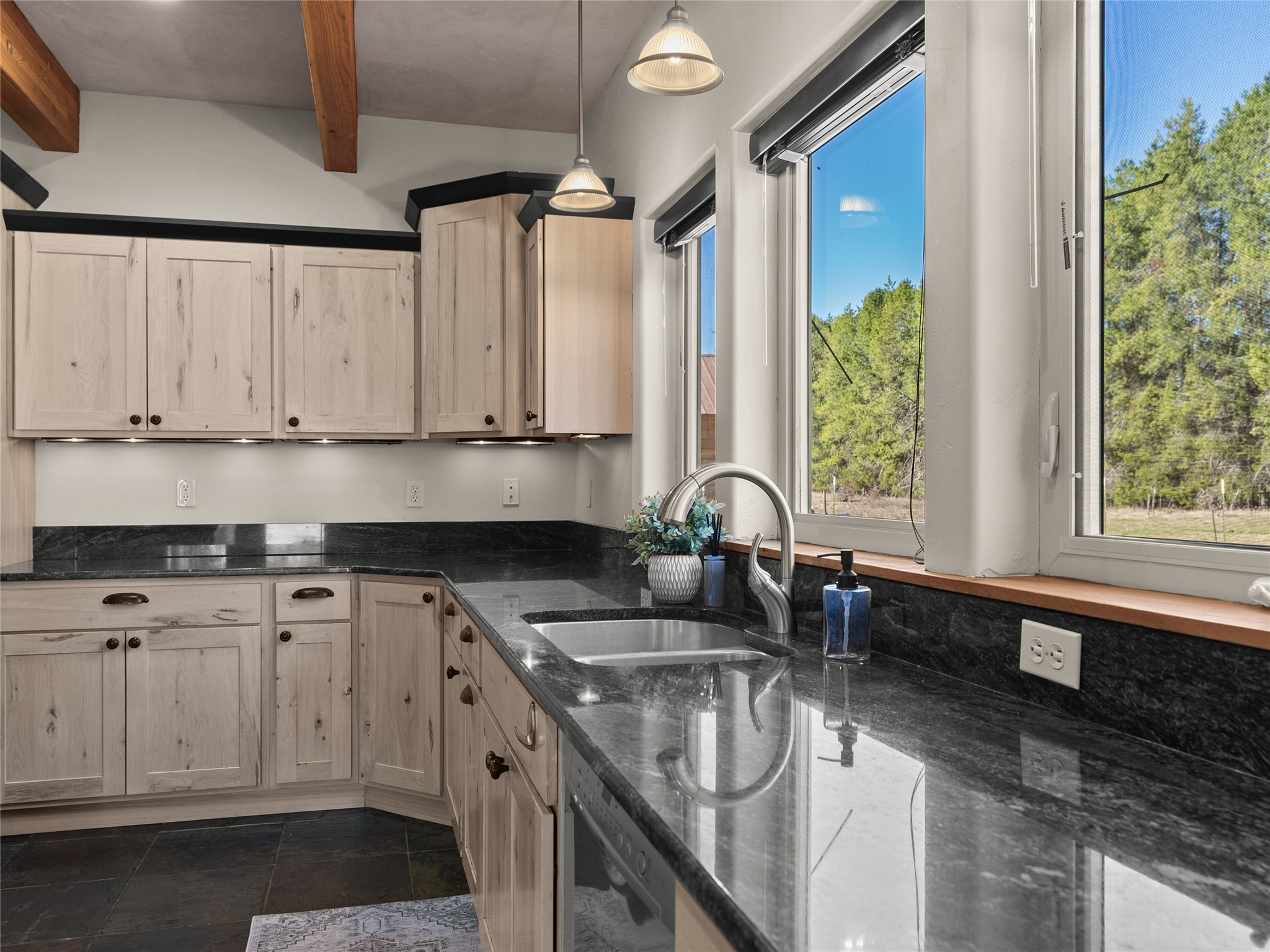 Kitchen with pale wood cabinets, black stone counters, a double sink, and large windows overlooking trees.