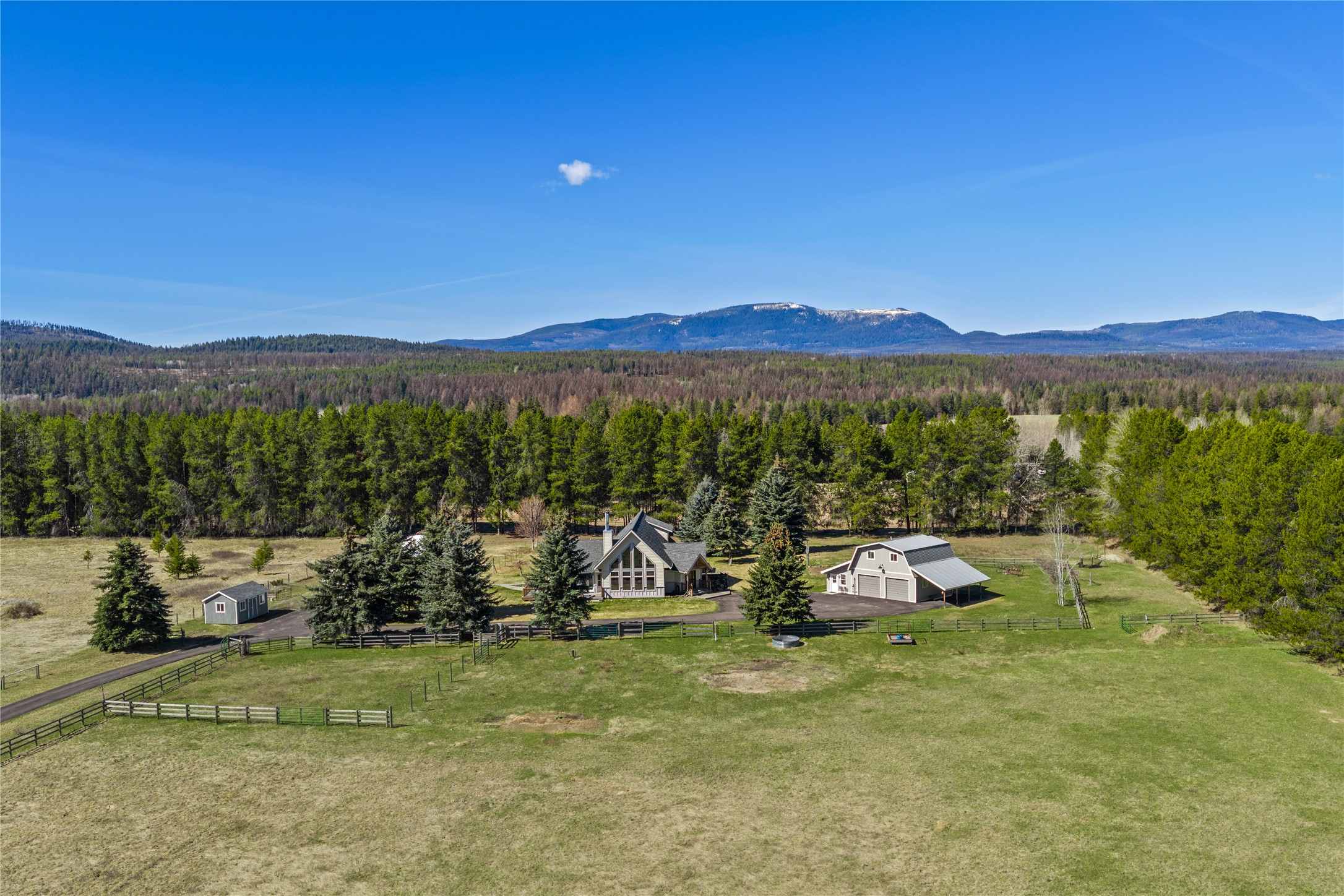 Aerial view of a home and barn amid fenced pasture, evergreen trees, and distant mountains.