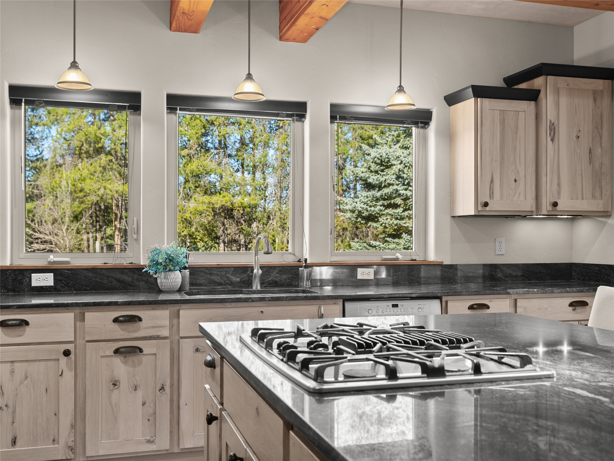 Kitchen with a gas cooktop on a large island and three windows overlooking trees.