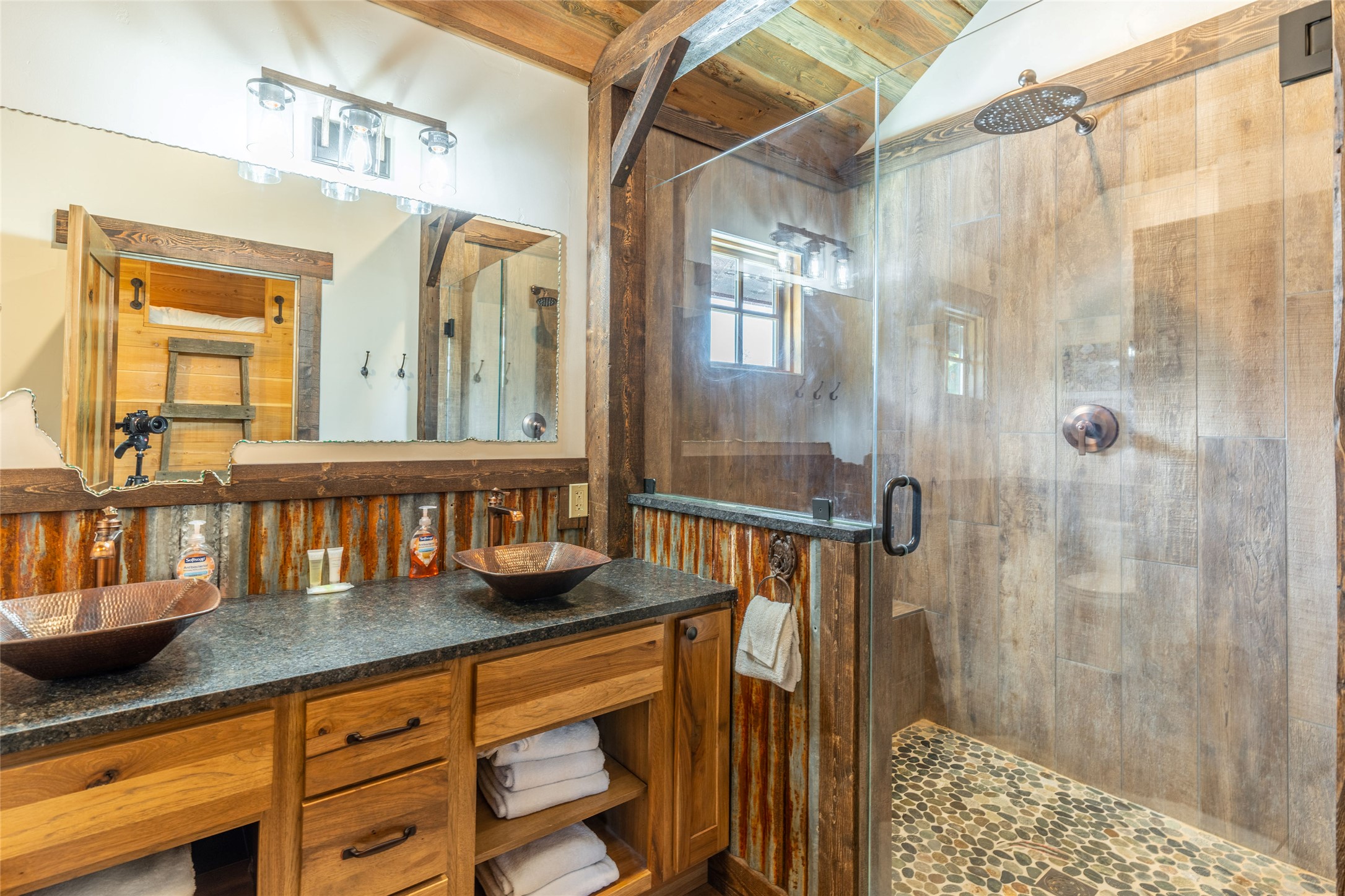 Rustic bathroom with copper vessel sinks and a glass walk-in shower with pebble flooring.