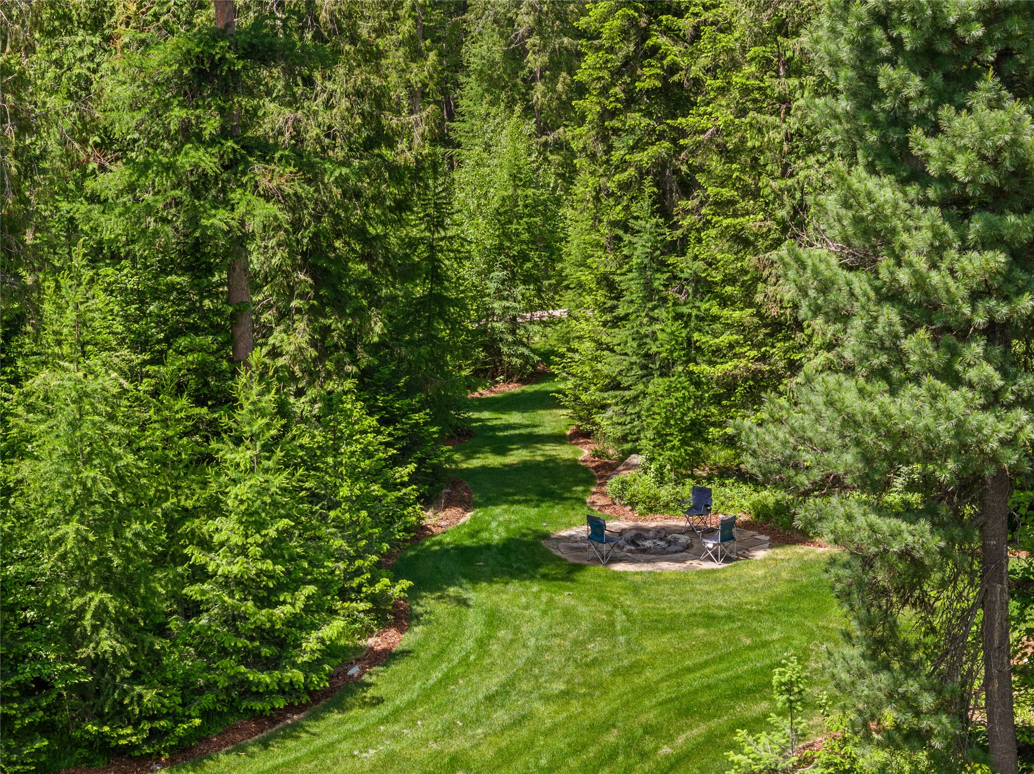 Wooded backyard lawn with a stone fire pit and three outdoor chairs.