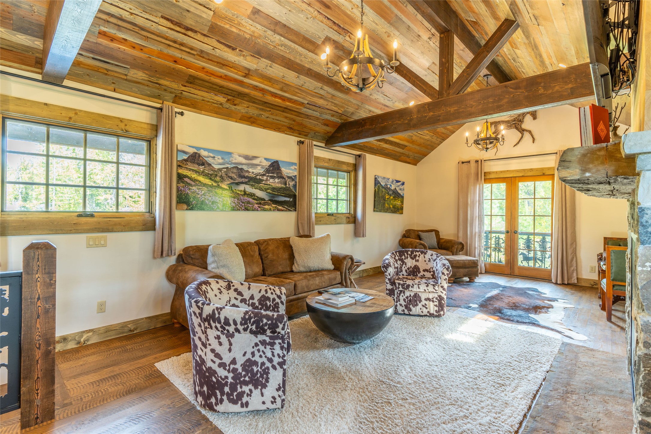 Rustic living room with exposed timber beams, cowhide-patterned chairs, and French doors opening to a balcony.