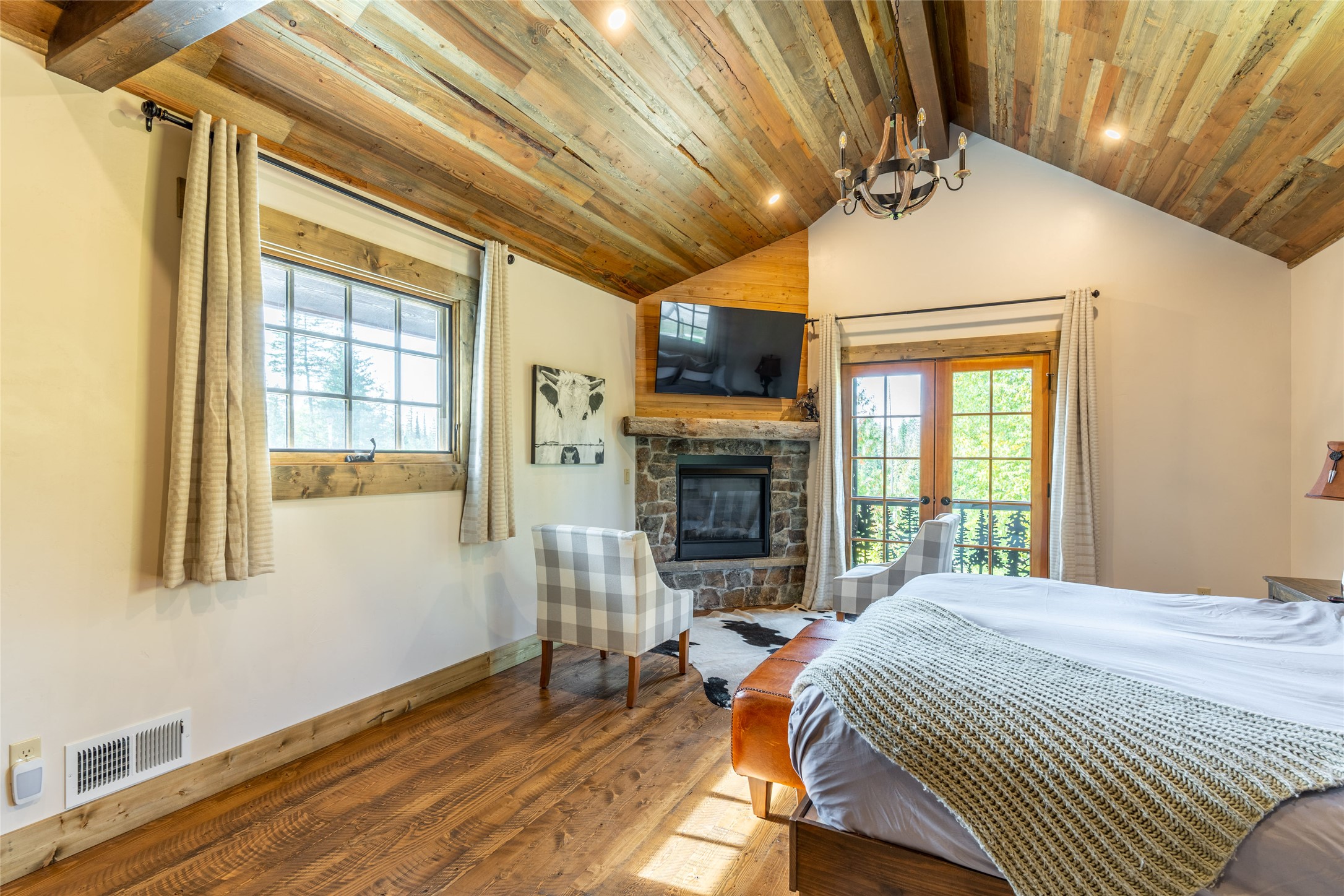 Bedroom with vaulted wood ceiling, stone fireplace, plaid chairs, and glass doors.