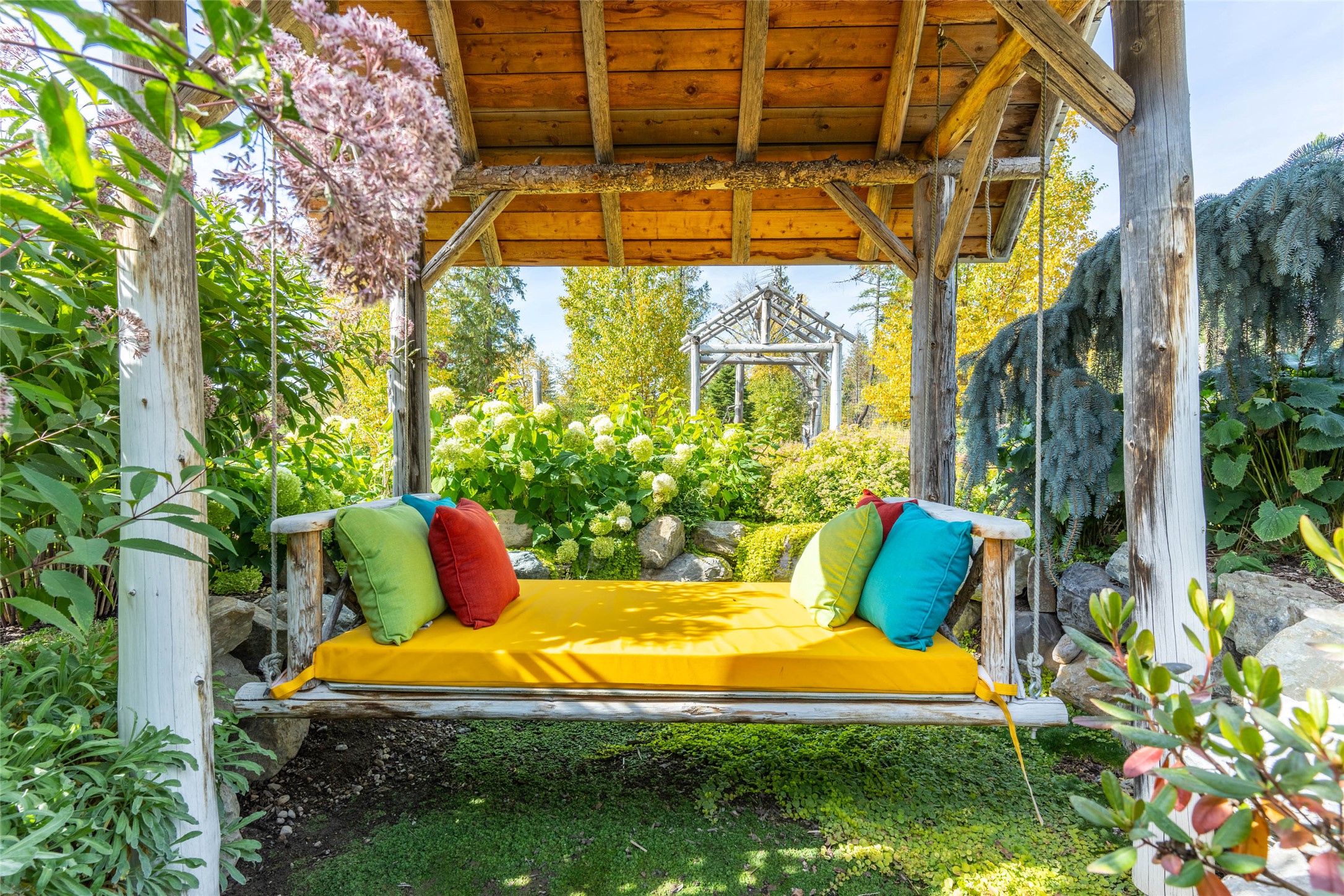 Garden swing beneath a timber pergola, with a yellow cushion and colorful pillows amid lush landscaping.