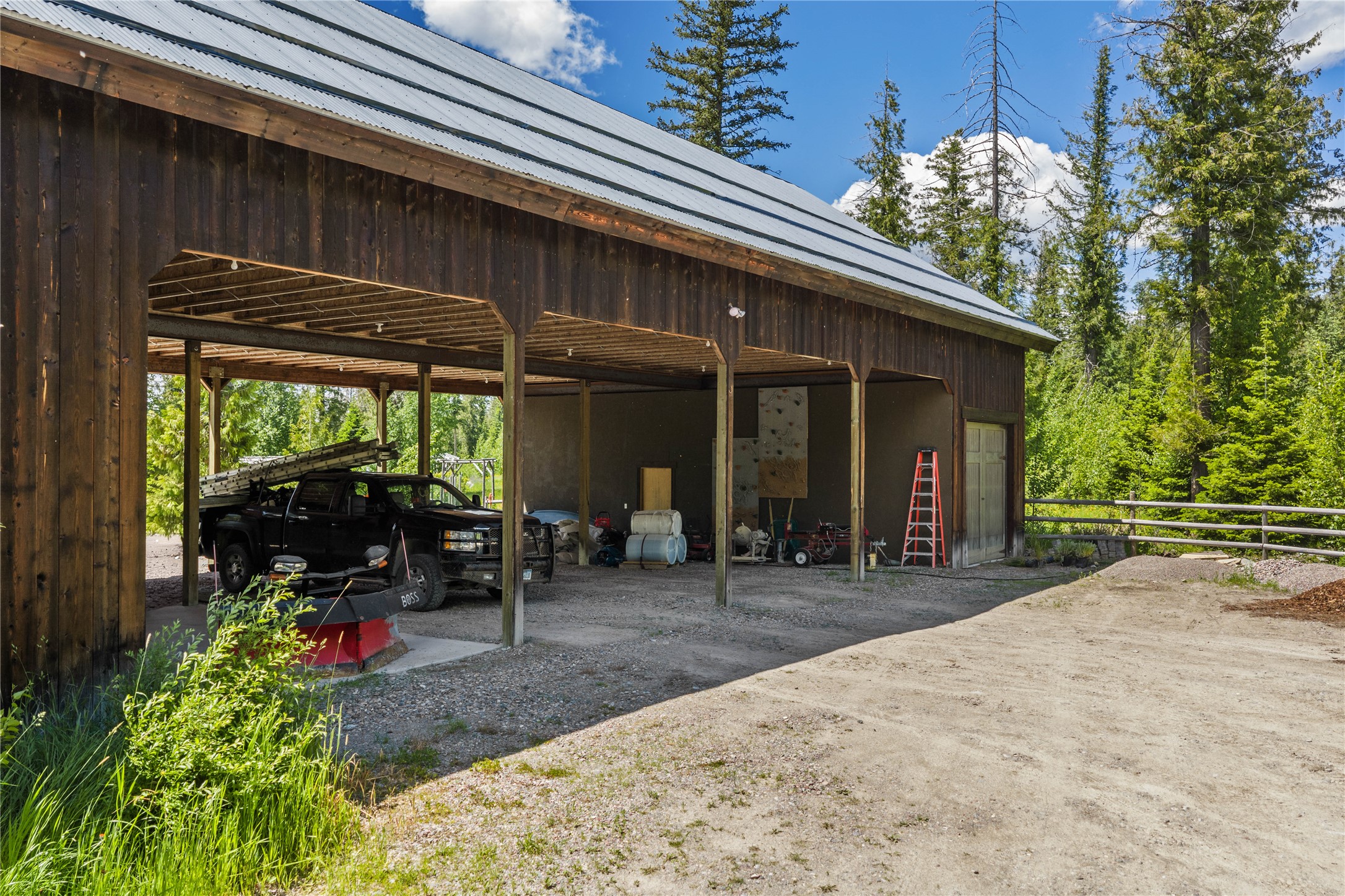 Timber barn and open equipment bays with a pickup truck, red ladder, and gravel drive.