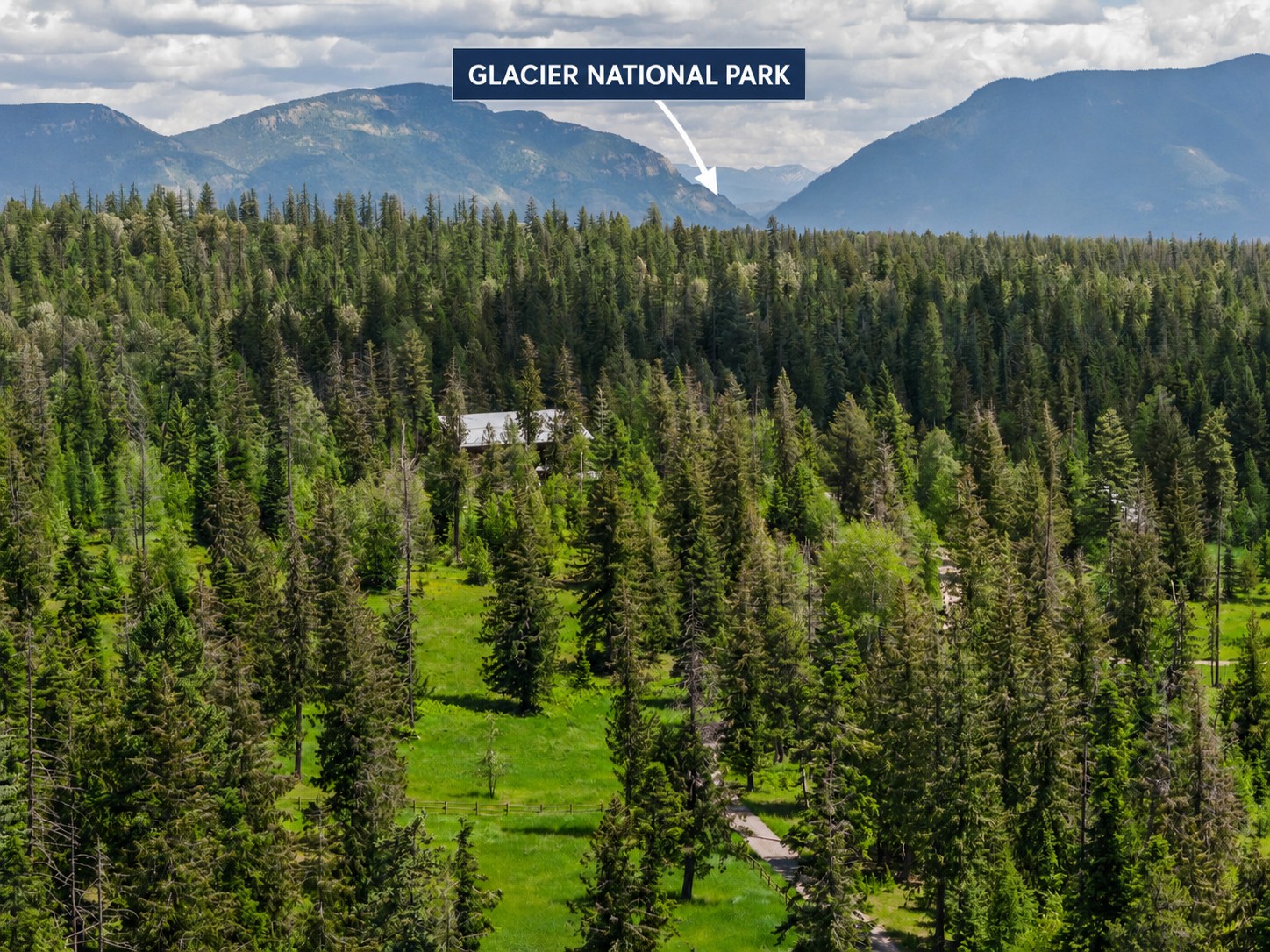 Aerial view of a wooded property with a cabin, meadow, and Glacier National Park beyond.
