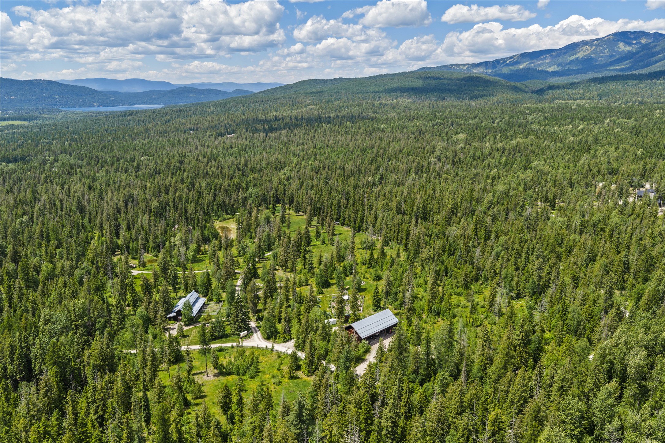 Aerial view of forested property with several buildings, mountain ridges, and a distant lake.