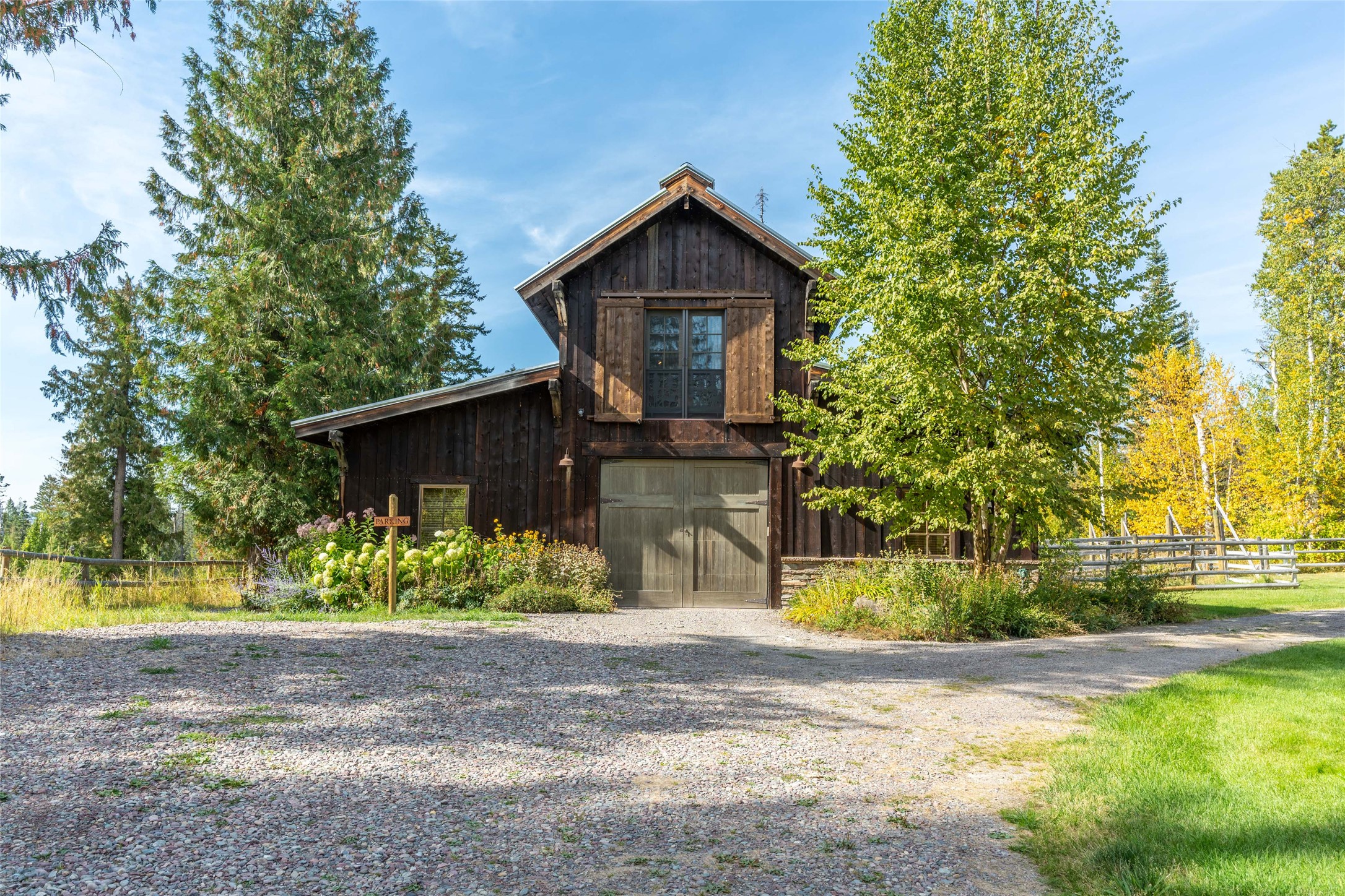 Dark timber two-story garage with a large double door, gravel driveway, and autumn trees.