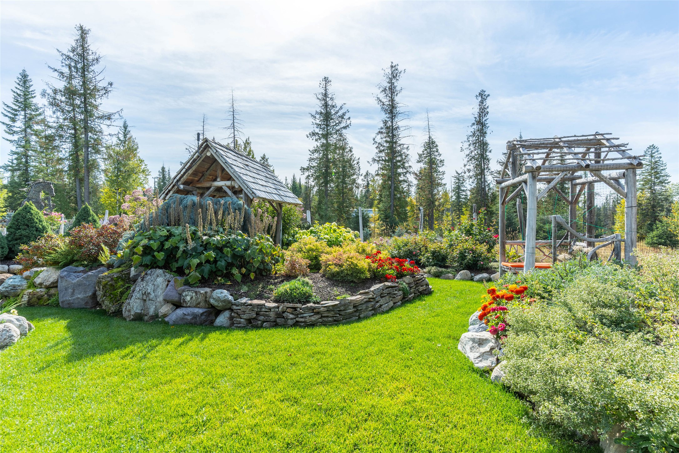 Landscaped garden with a timber shelter, colorful flower beds, stone walls, and a rustic wooden arbor.