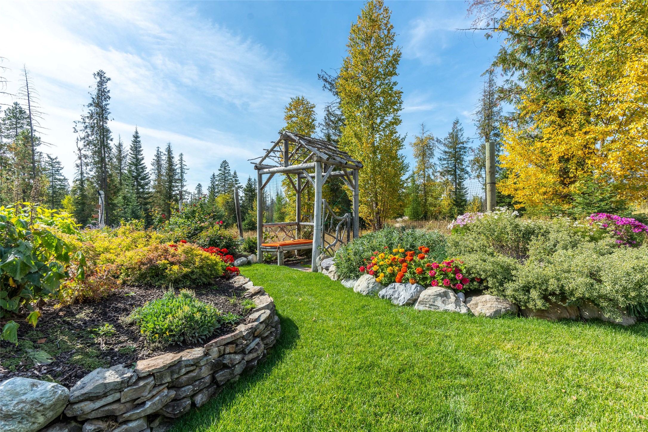 Landscaped garden with colorful flower beds, curved stone walls, and a rustic timber arbor.