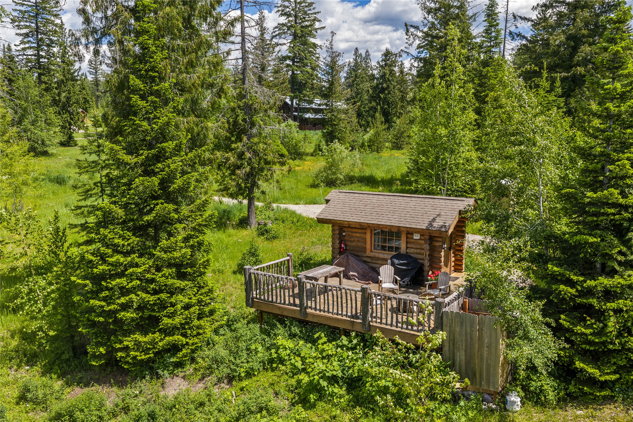 Log cabin with a furnished wooden deck surrounded by trees and a grassy clearing.