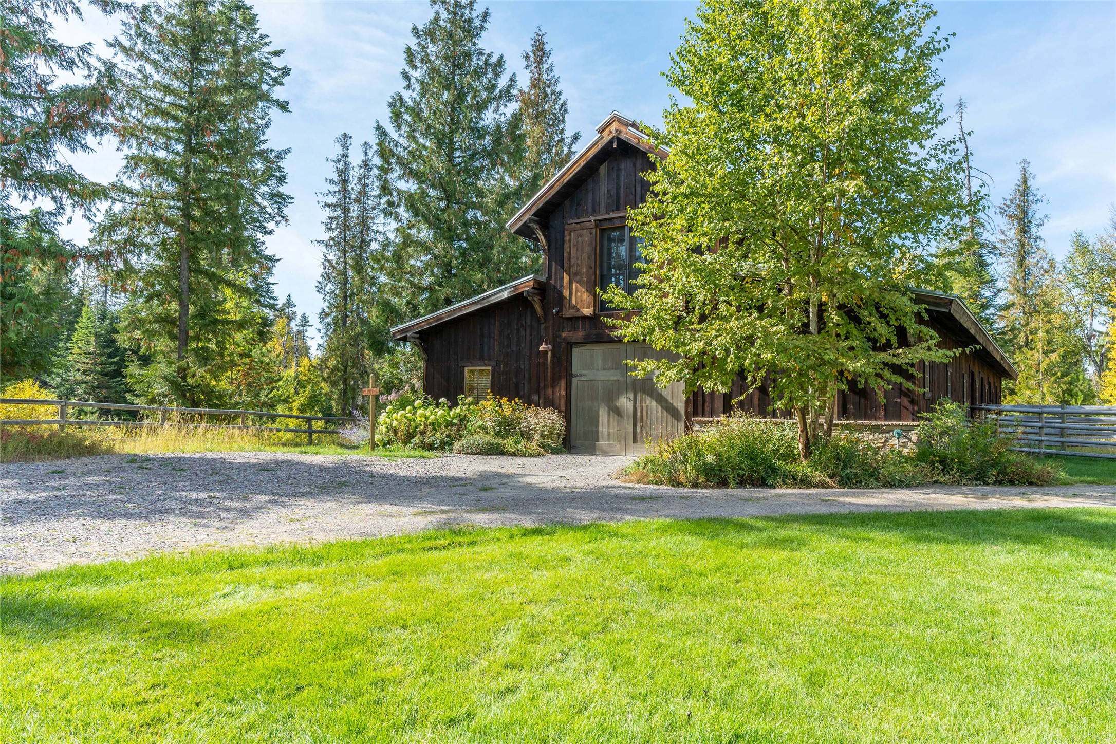 Dark wood home with a tall gable, gravel drive, garden, and lawn among evergreen trees.