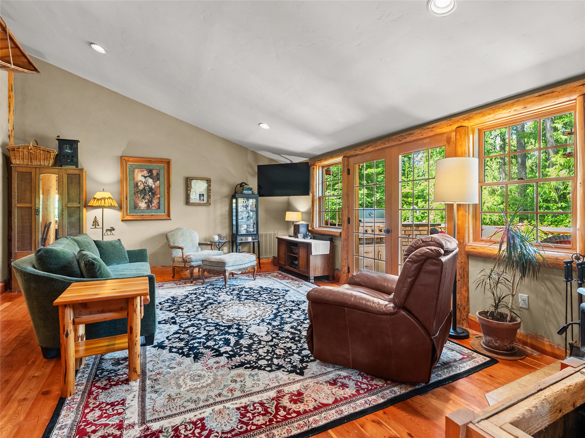 Living room with wood-trimmed windows overlooking trees, hardwood floors, and a large patterned rug.