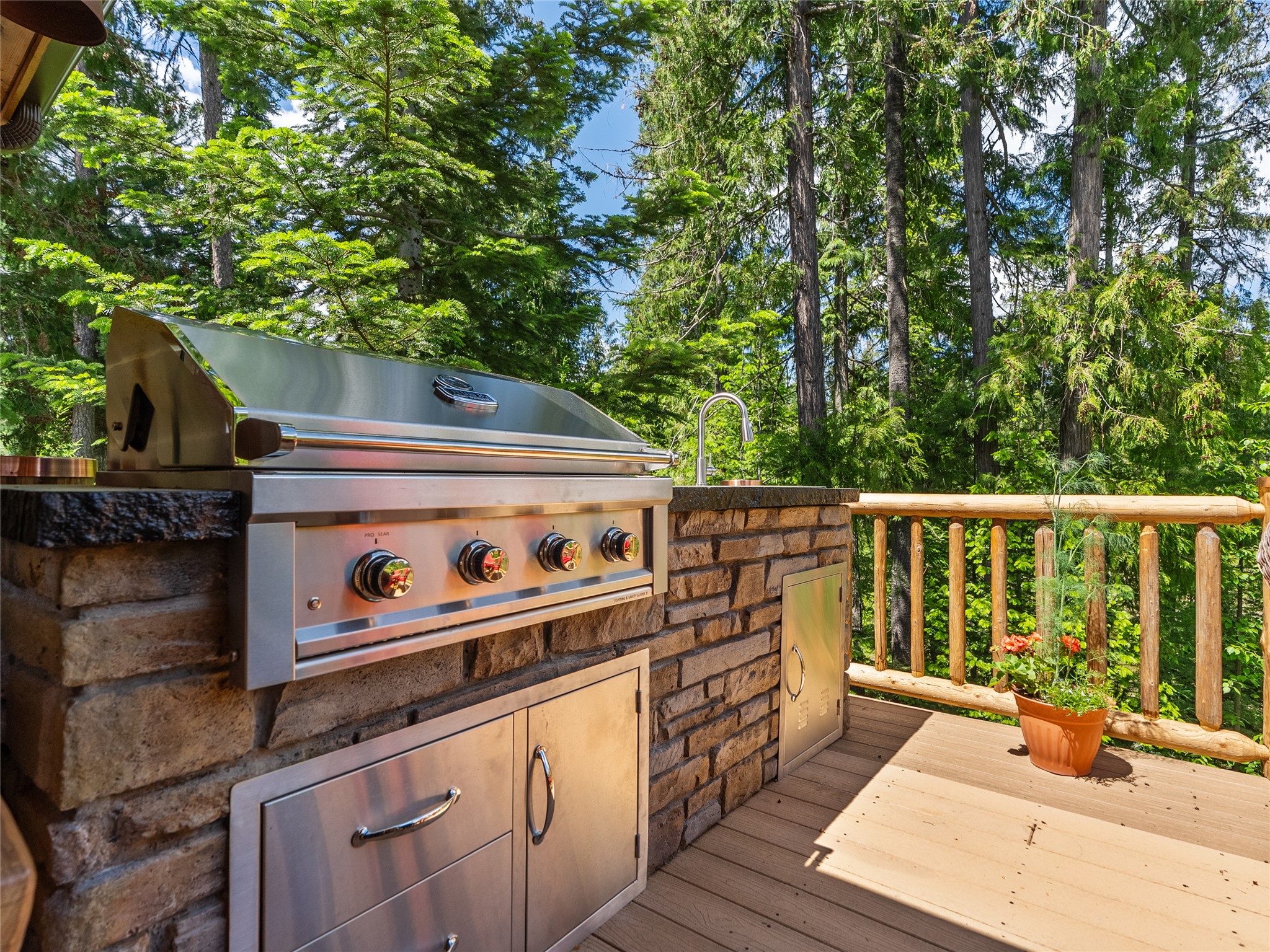 Outdoor deck kitchen with a stainless steel grill and sink beside a wooded view.