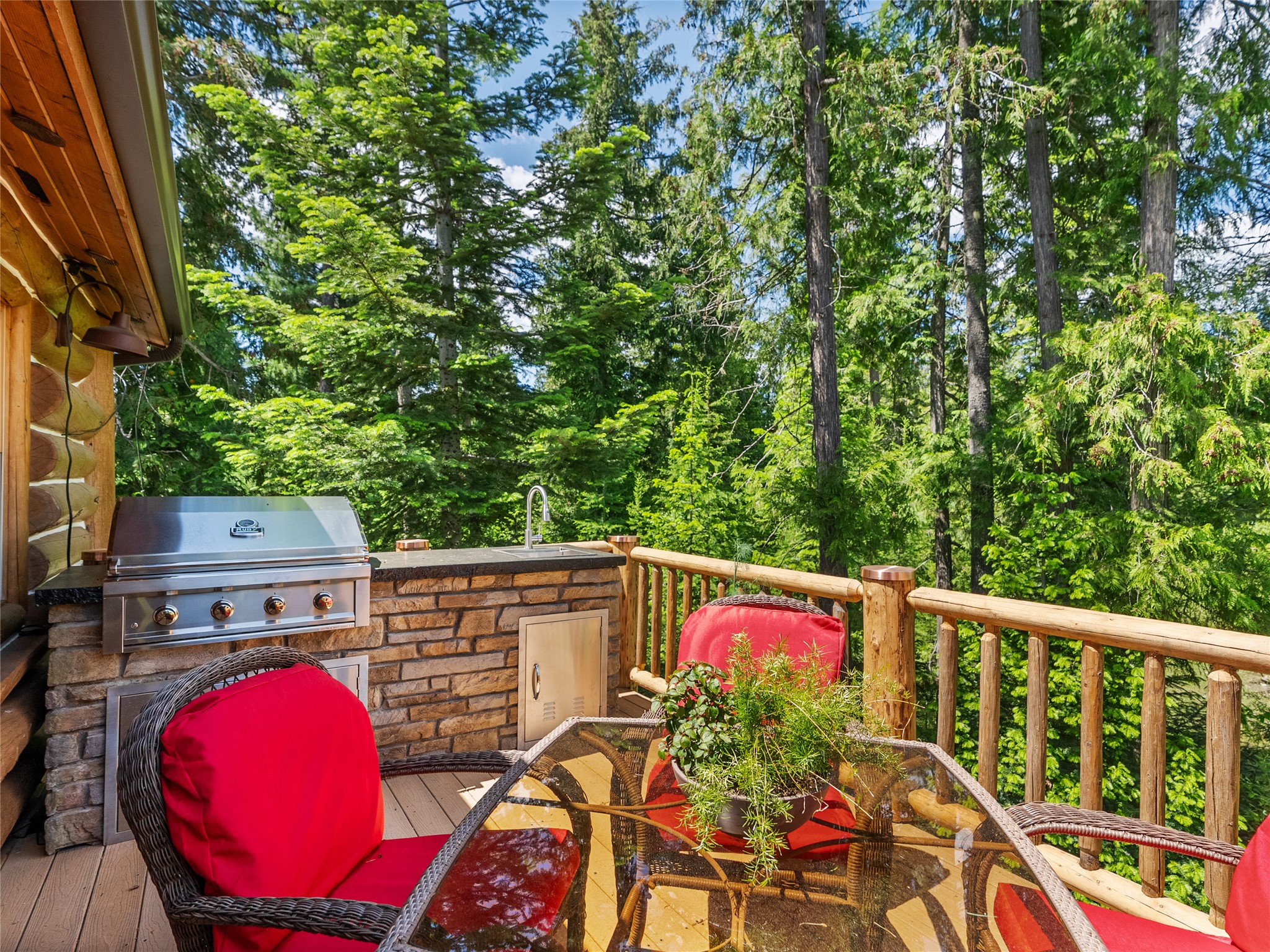 Outdoor deck with a built-in grill and sink, red-cushioned chairs, and a view of evergreen forest.