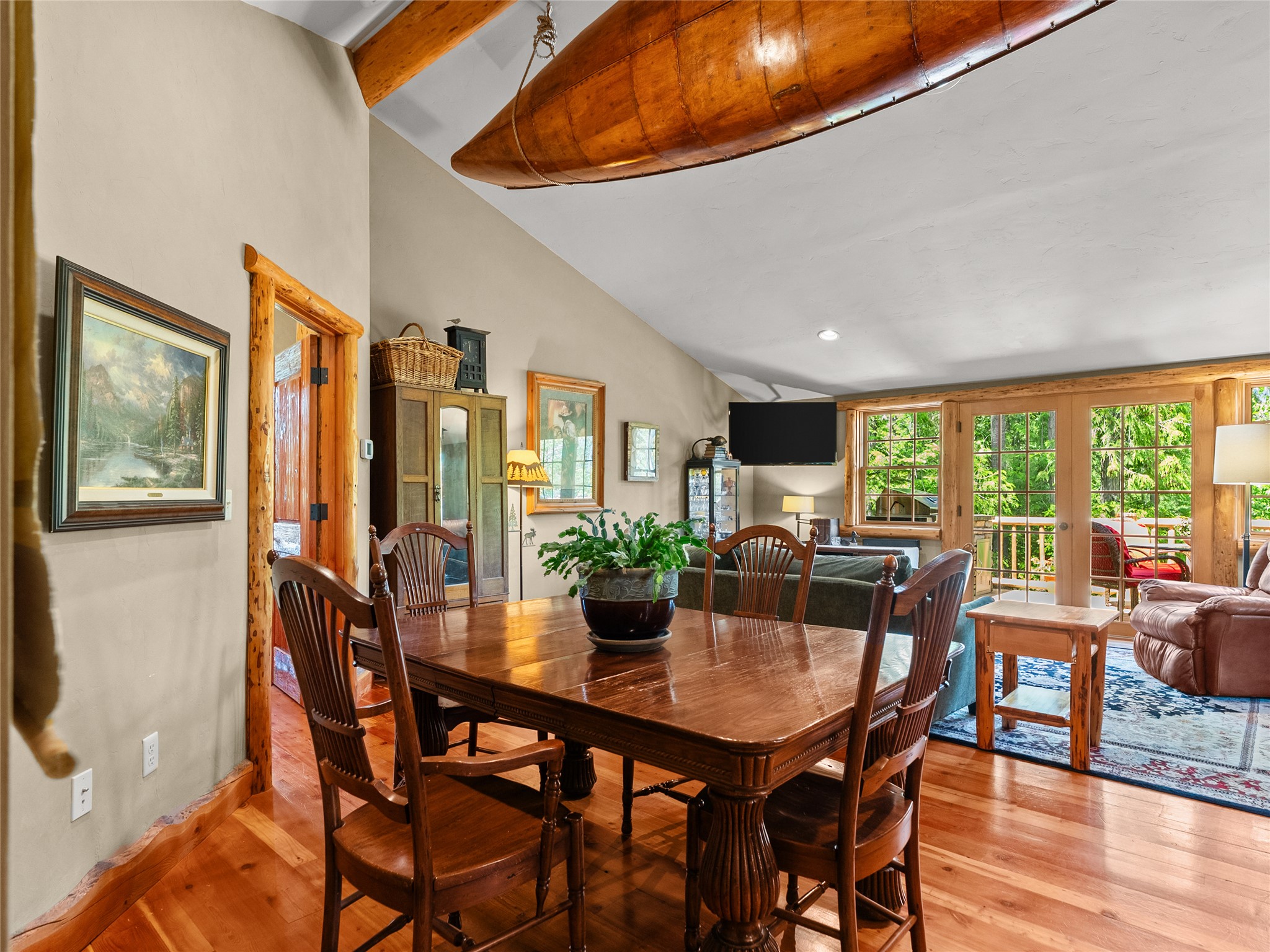 Dining and living area with a polished wood table, ceiling-mounted canoe, and windows overlooking greenery.