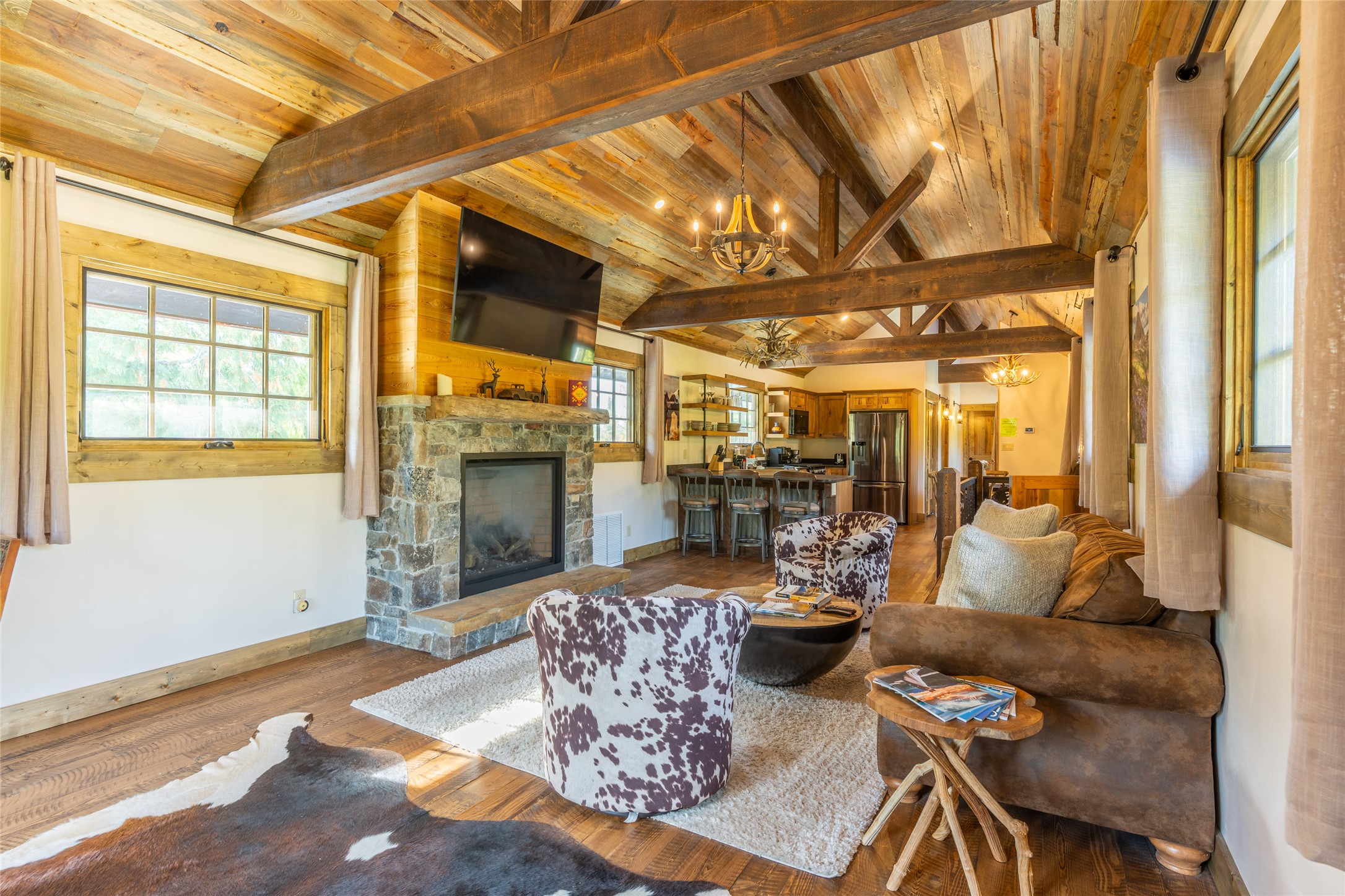 Rustic living room with vaulted timber ceiling, stone fireplace, and cowhide-patterned chairs.