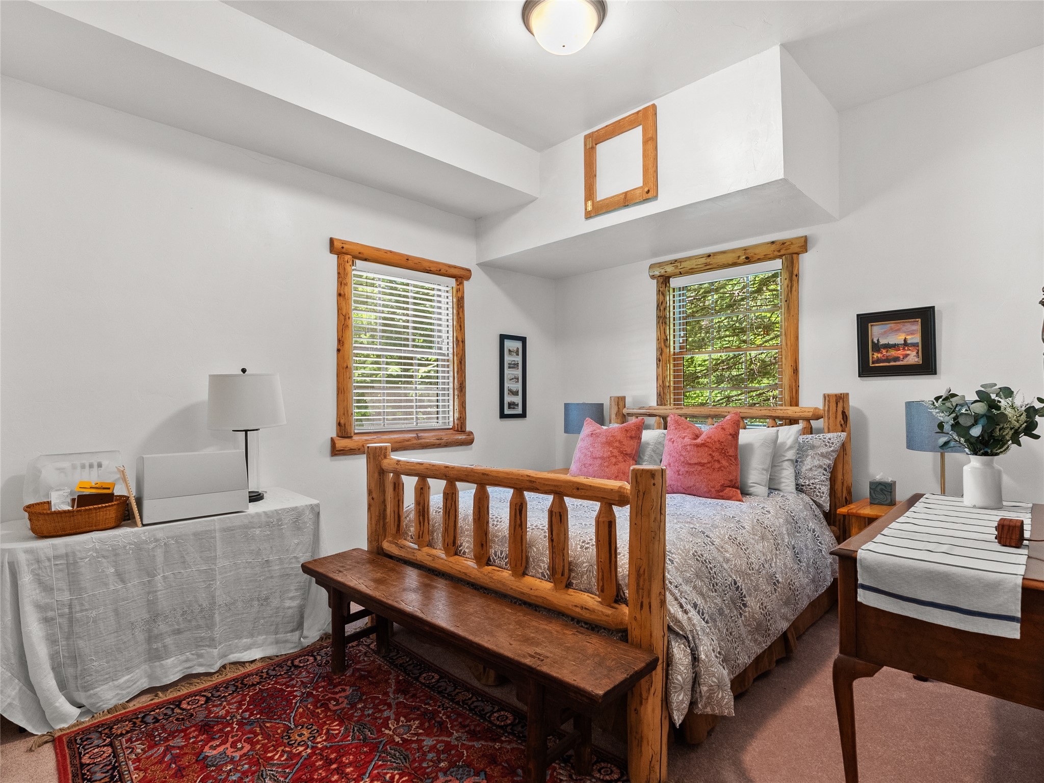 Bedroom with a rustic log-frame bed, pink pillows, wood-trimmed windows, and a red patterned rug.