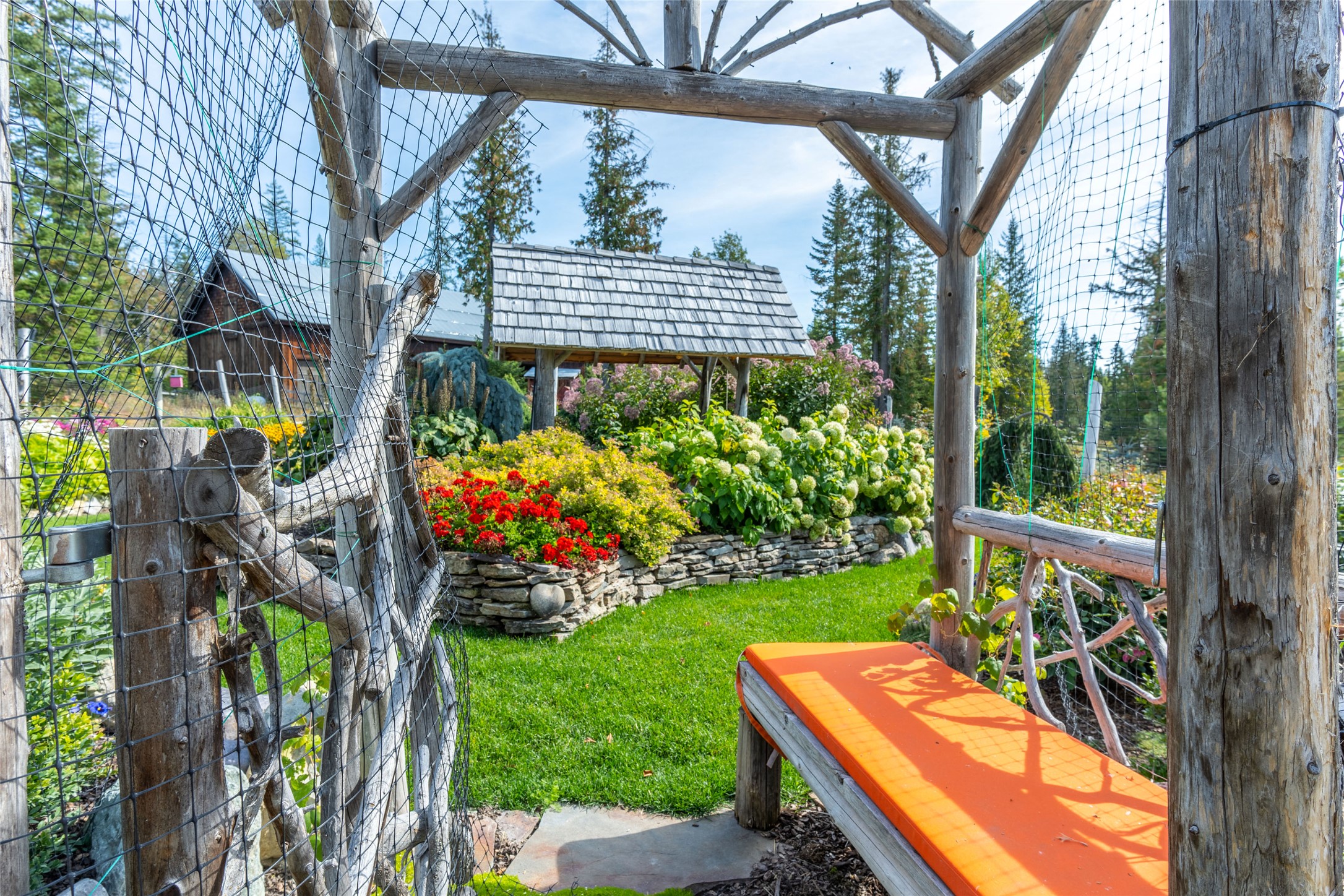 Landscaped garden with a timber pergola, orange-cushioned bench, colorful raised flower beds, and lawn.