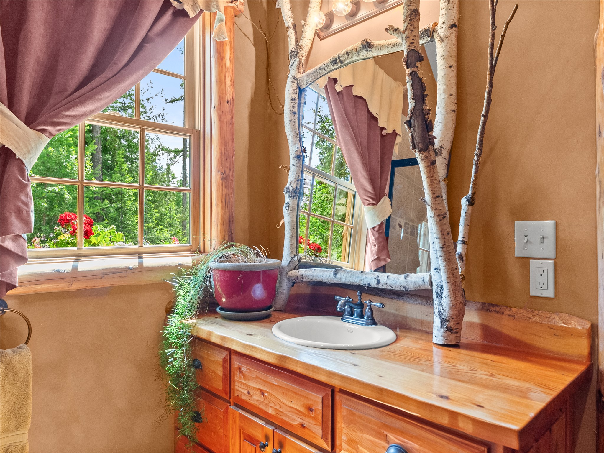 Bathroom vanity with a birch-branch mirror, live-edge counter, and window with mauve curtains.