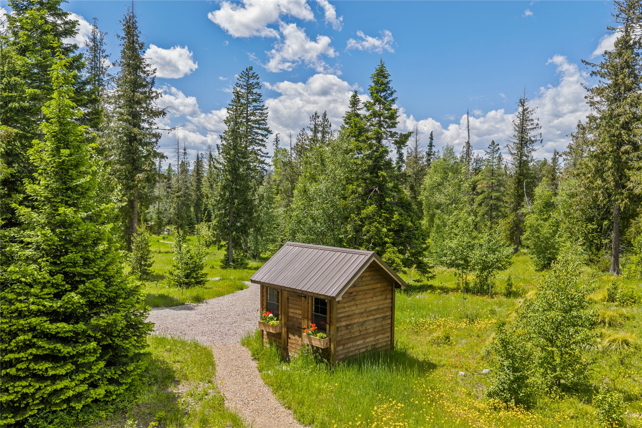 Small wooden cabin with flower boxes beside a gravel path in a wooded clearing.