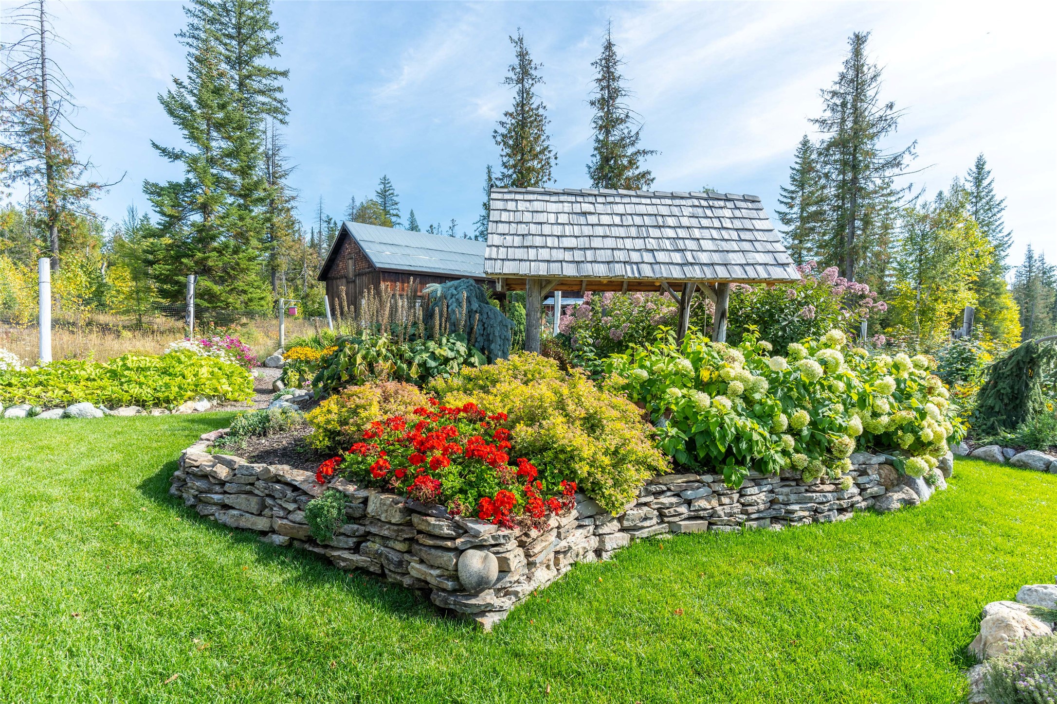 Landscaped garden with colorful flower beds, a stone retaining wall, and a timber shelter among evergreens.