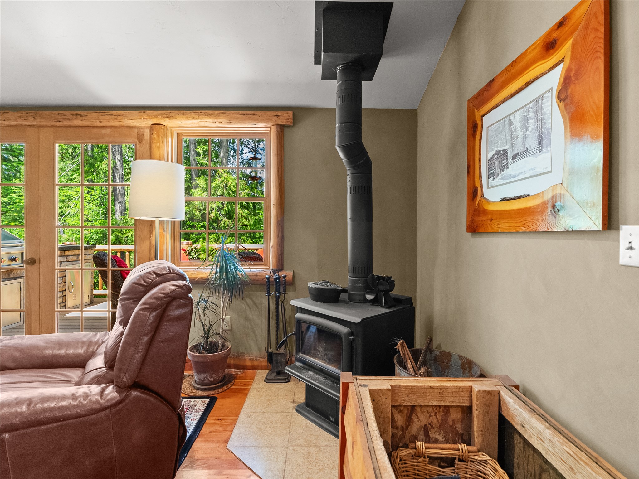 Living room with a black wood stove, brown leather recliner, and wood-trimmed windows overlooking green trees.