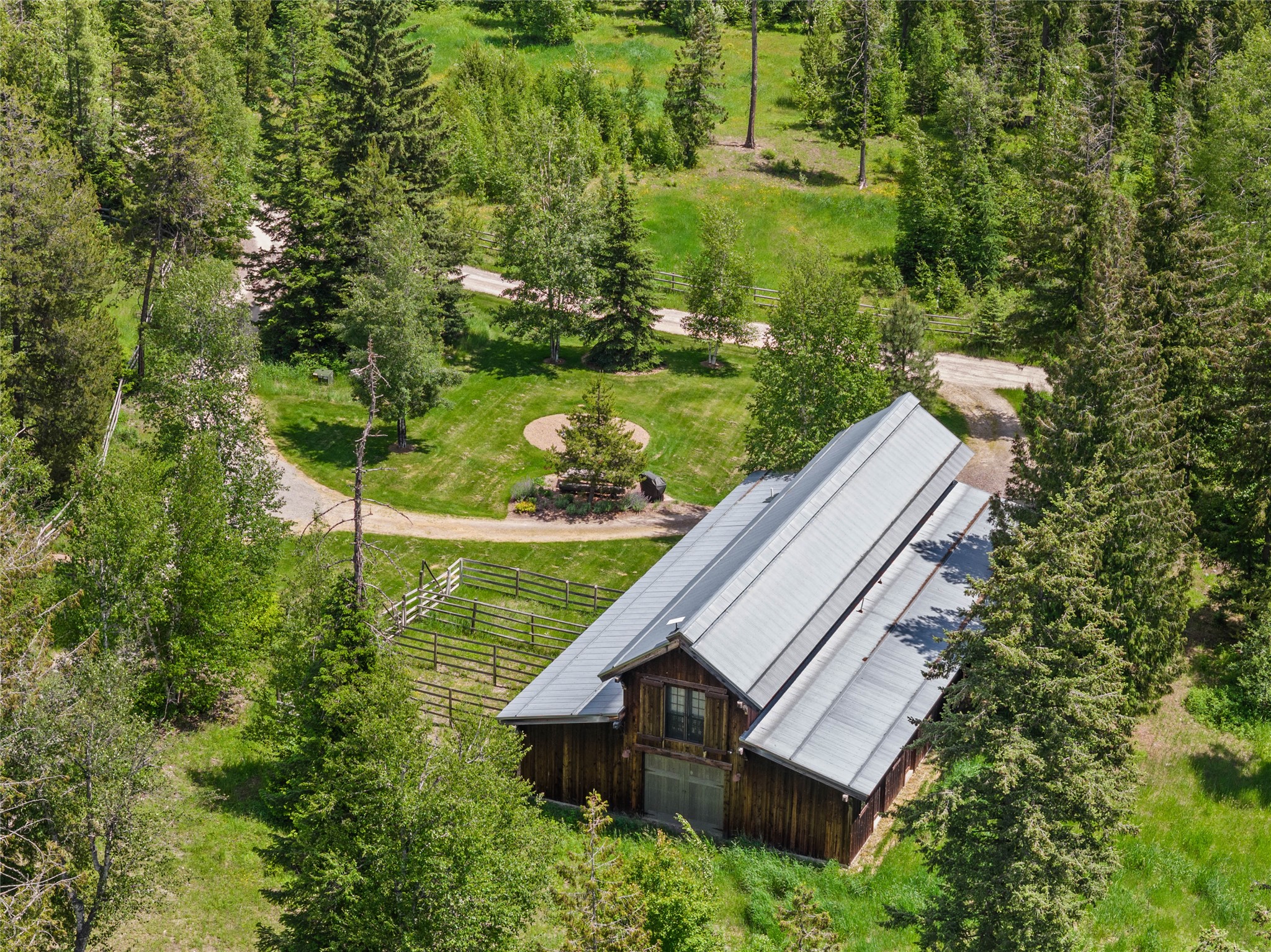 Aerial view of a timber home with a silver roof among trees, lawn, and fenced areas.