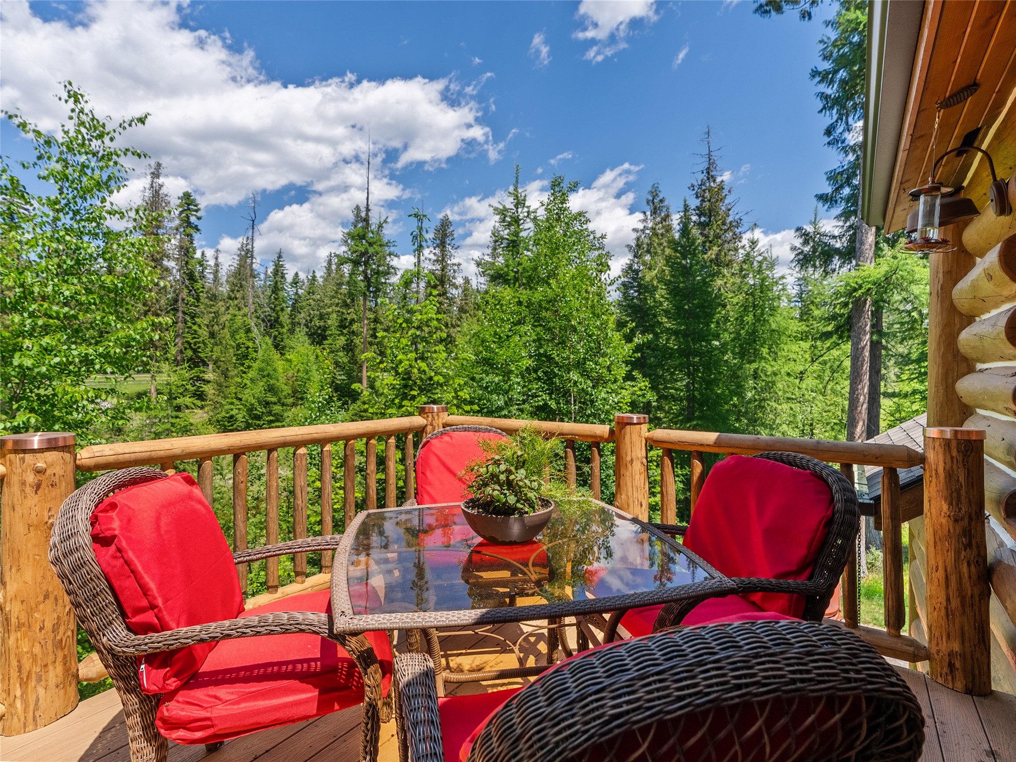 Log home deck with red-cushioned wicker chairs and a glass table overlooking a forest.