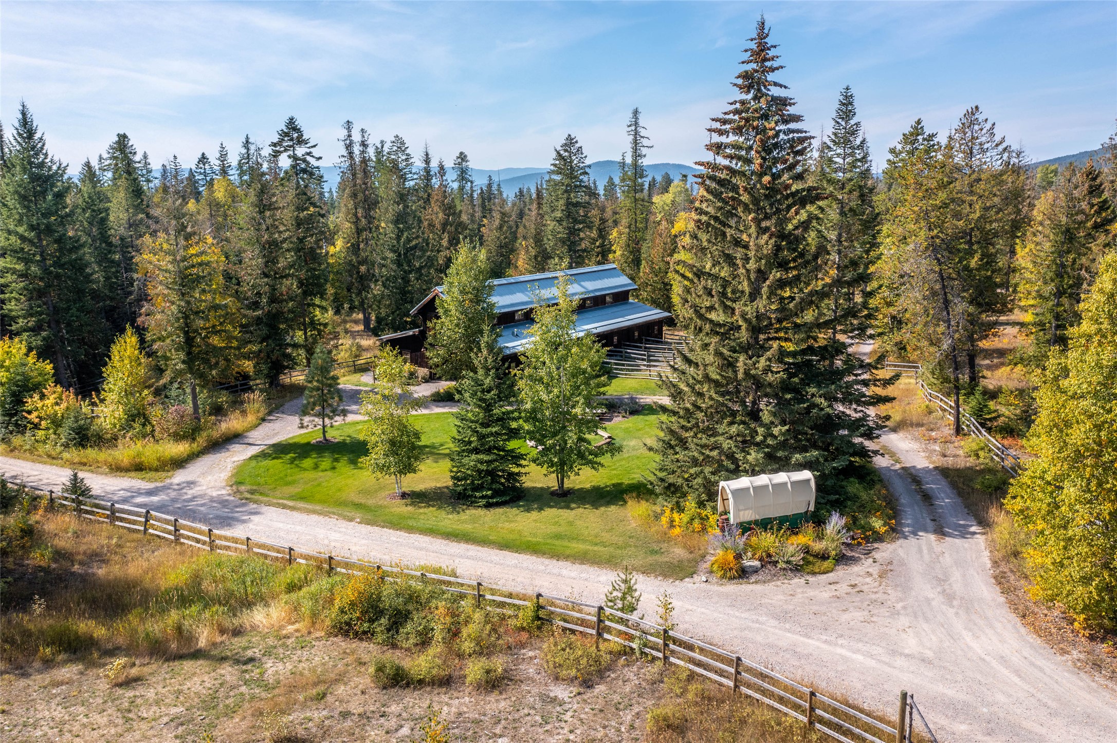 Aerial view of a timber home amid autumn woods, with a looping gravel drive and distant mountain ridges.