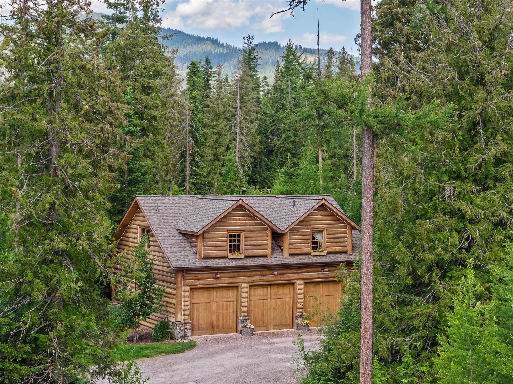 Log home with three garage doors nestled among evergreens, with a forested mountain behind it.