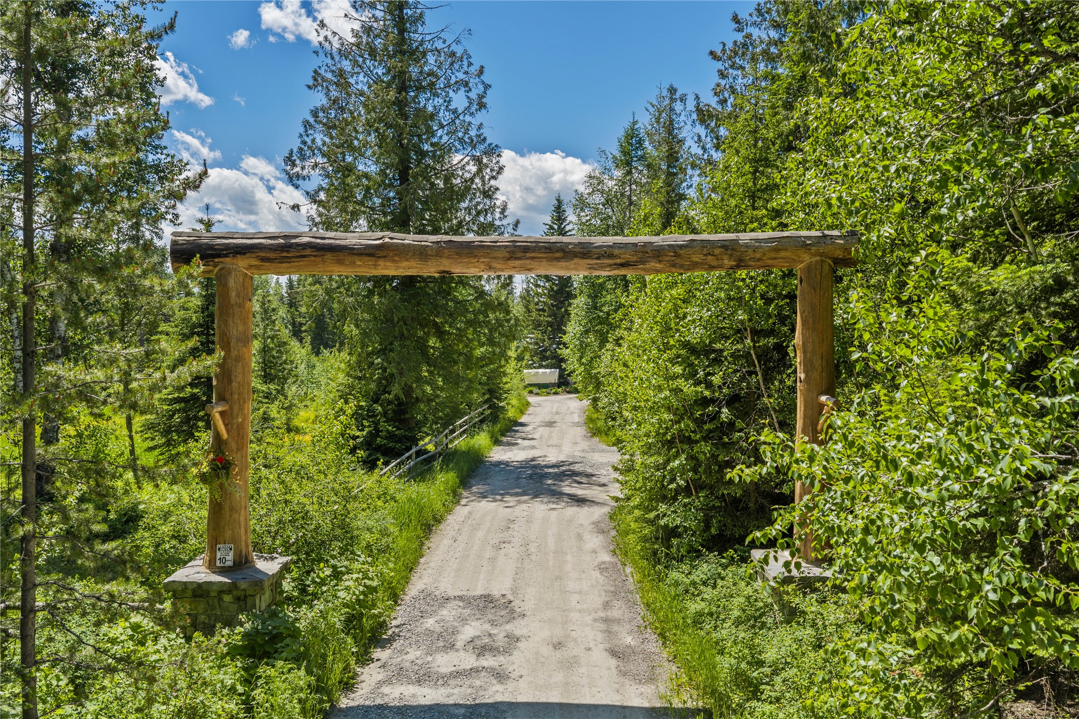 Gravel driveway beneath a rustic timber entrance arch, surrounded by dense green trees.
