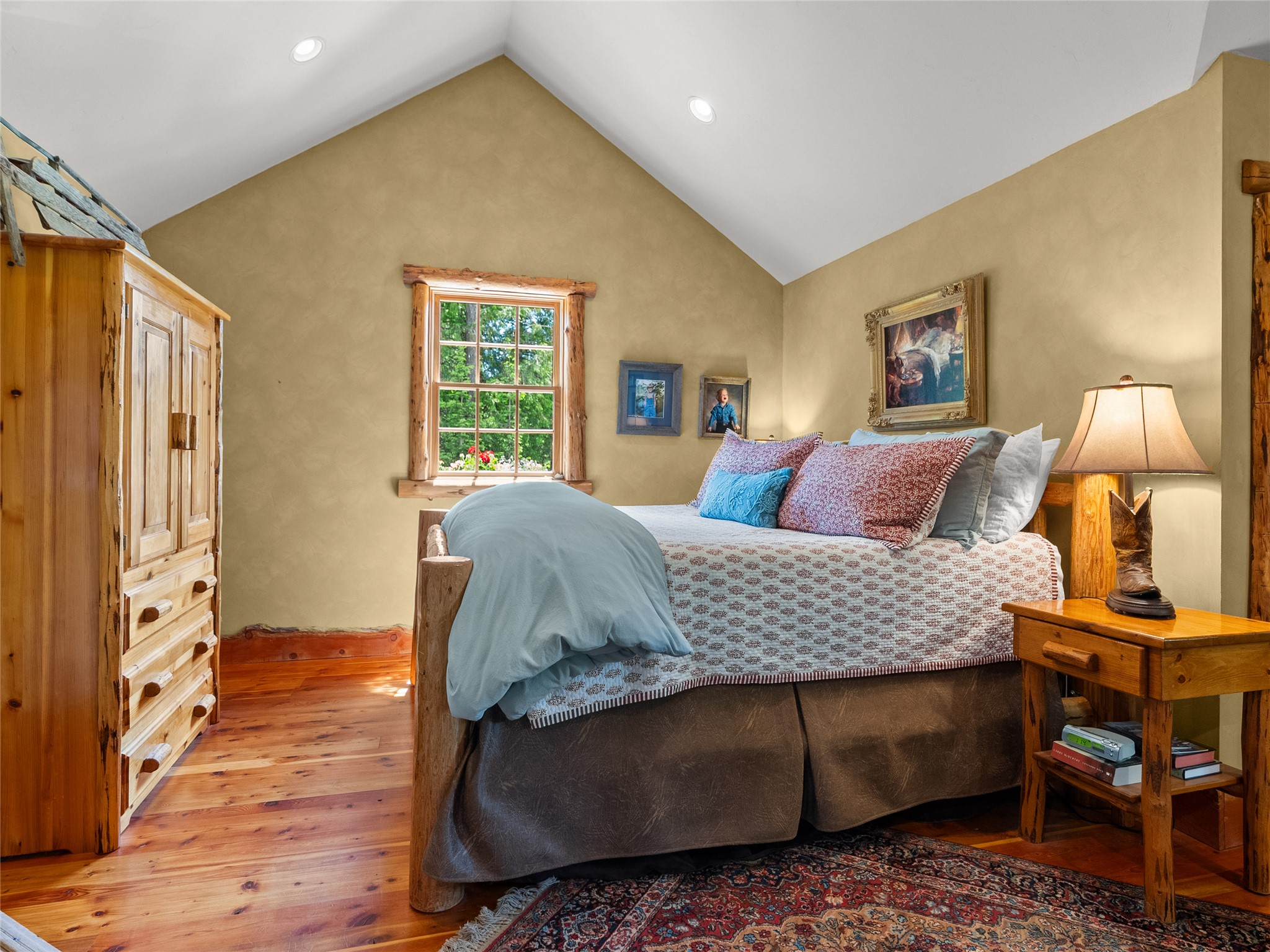 Bedroom with a vaulted ceiling, log-trimmed window, wood floors, and a large bed with blue and patterned bedding.