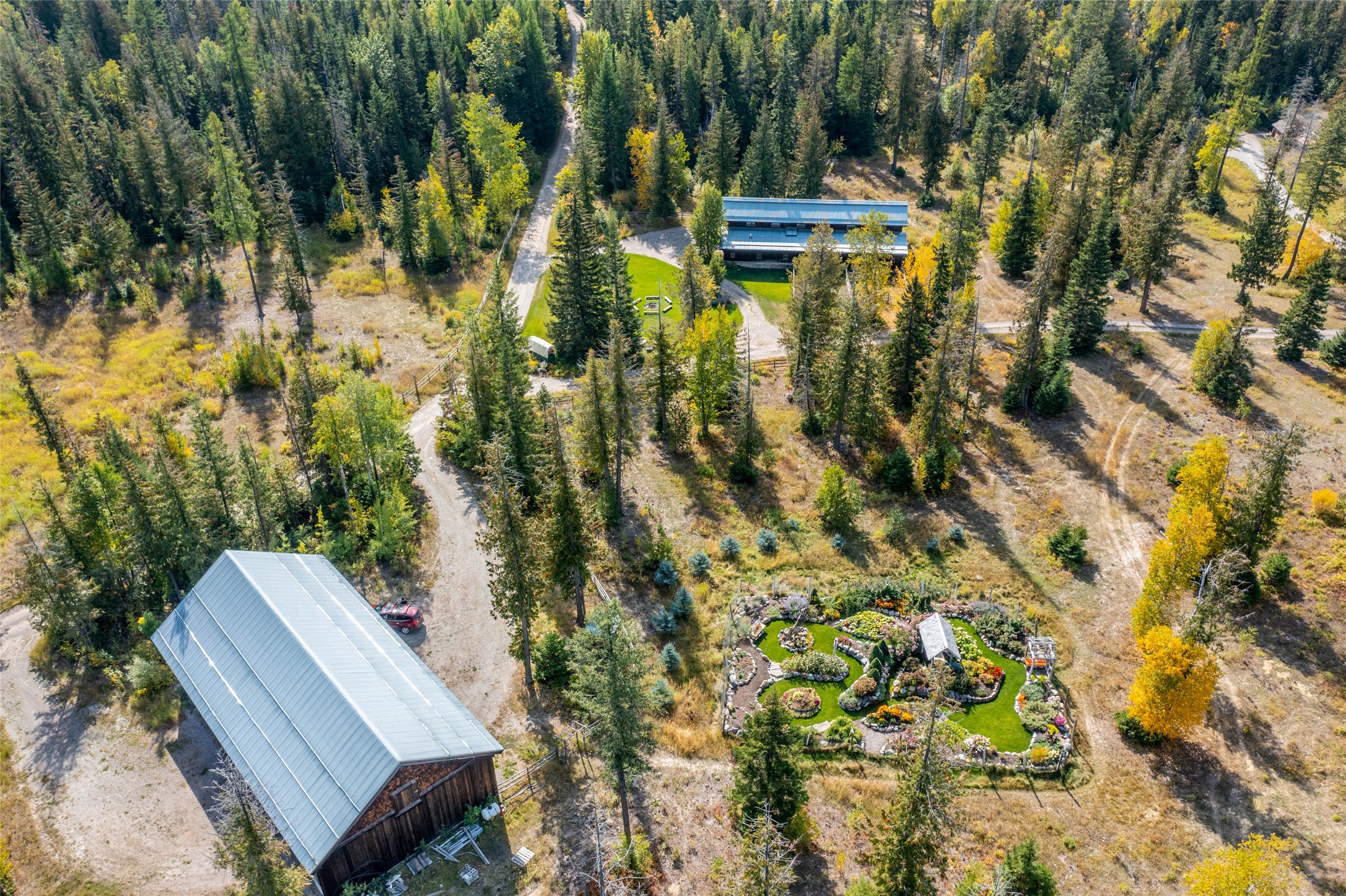 Aerial view of wooded property with two metal-roofed buildings and a colorful landscaped garden.