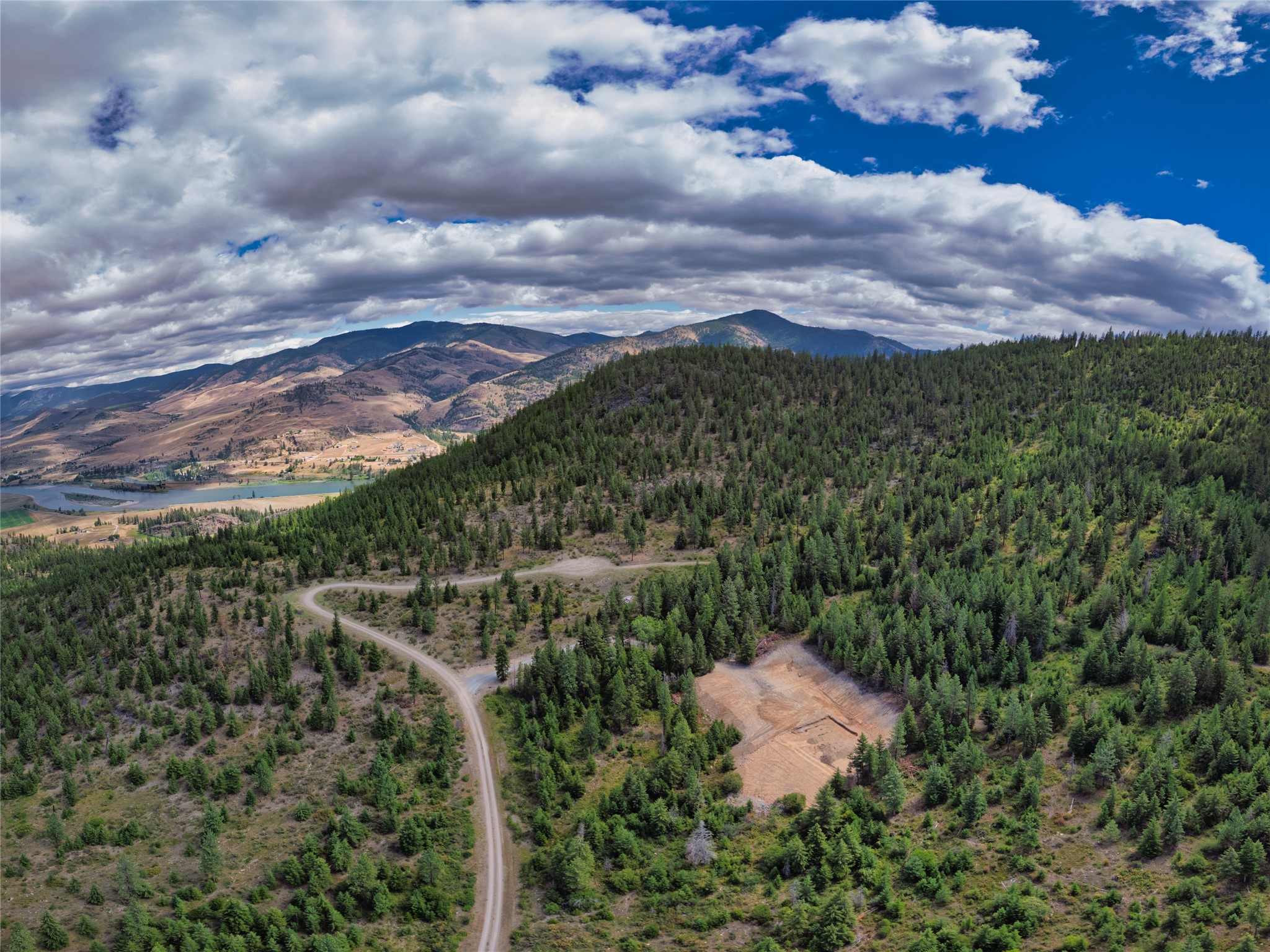 Aerial view of a wooded Montana hillside with a winding road, cleared site, river valley, and distant mountains.