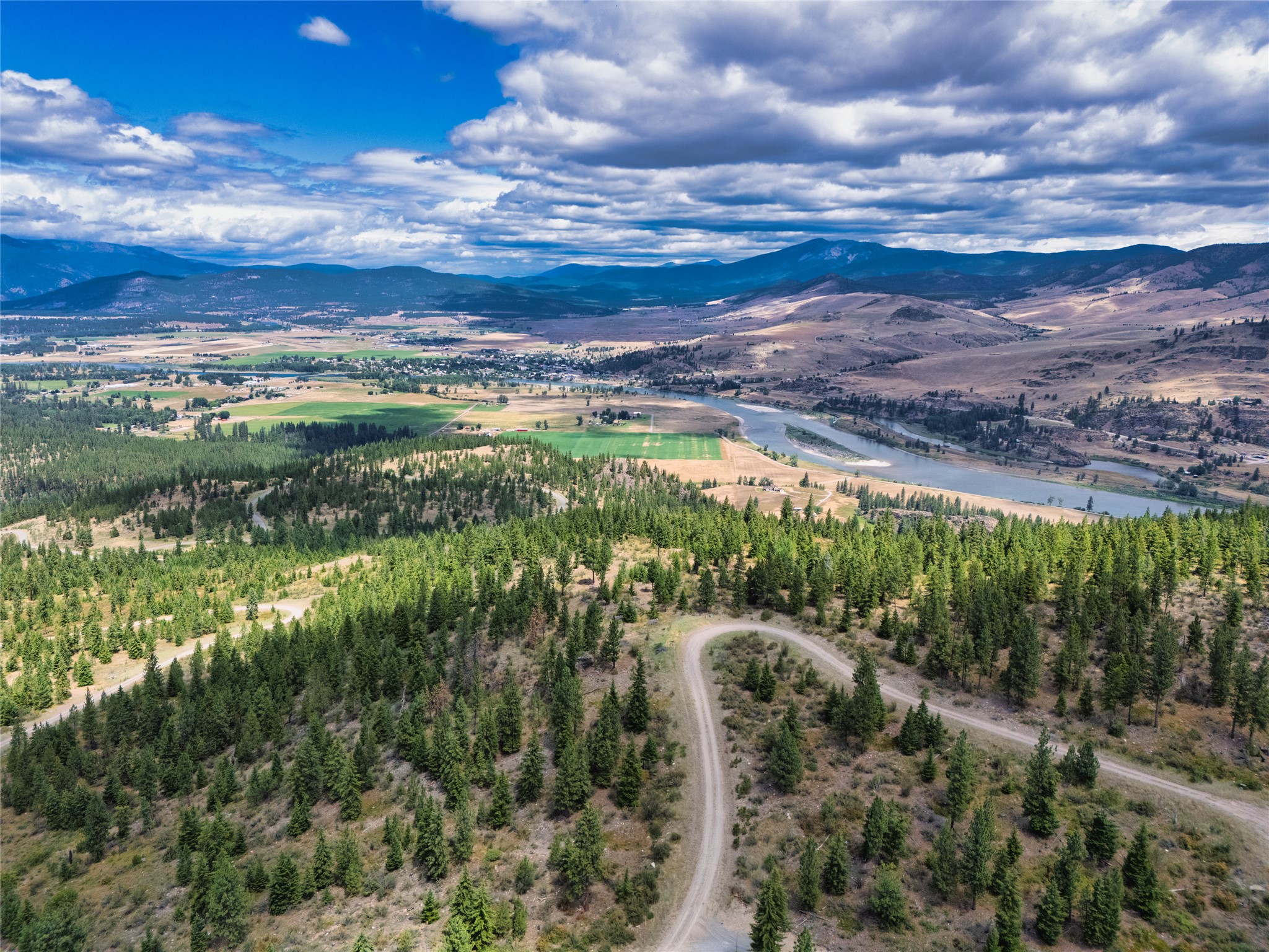 Aerial view of wooded land and a winding dirt road above a river valley and surrounding mountains.