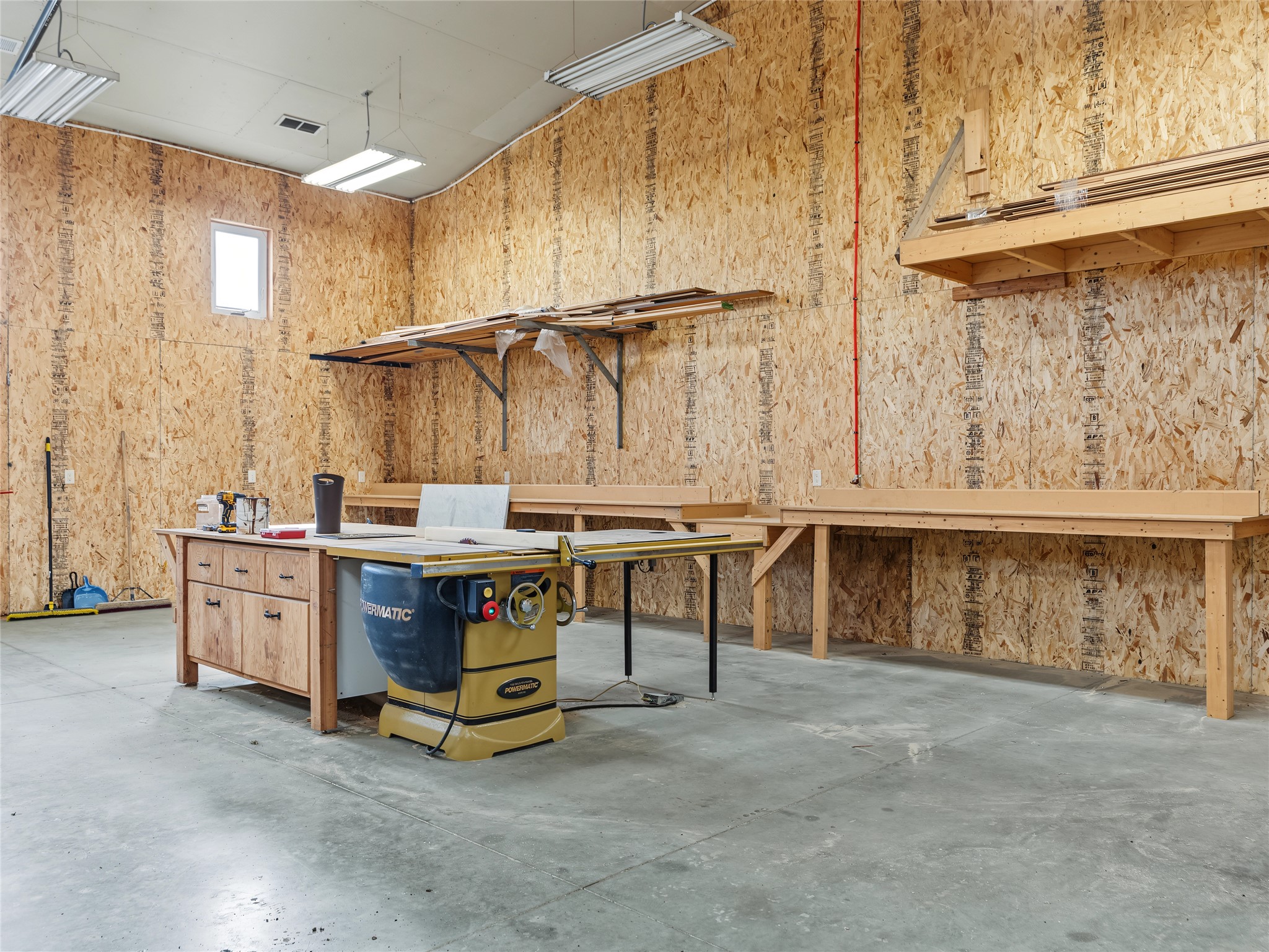 Woodworking workshop with a large table saw, long workbenches, and exposed OSB walls.