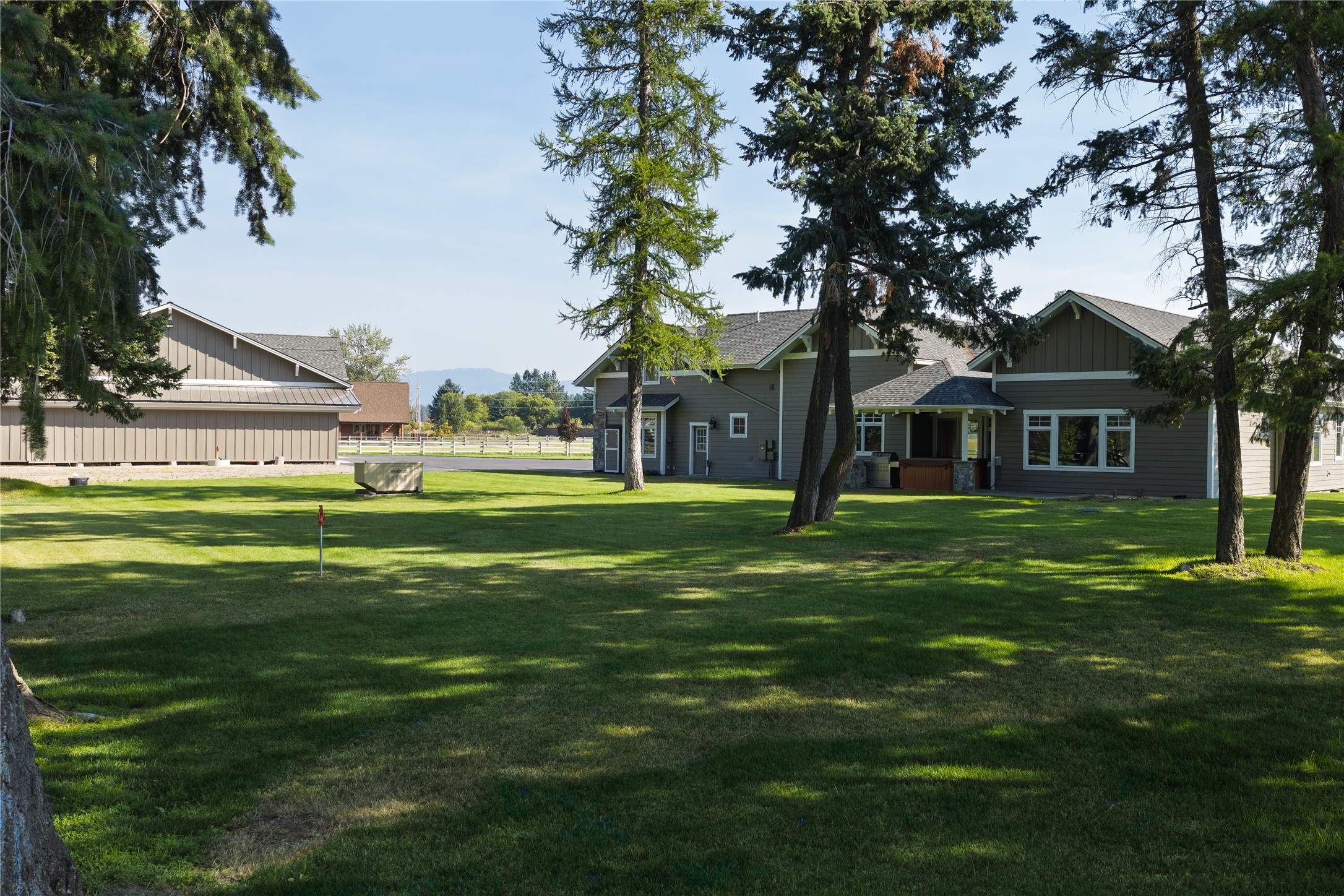 Gray home and detached outbuilding beyond a shaded lawn, framed by tall evergreens.