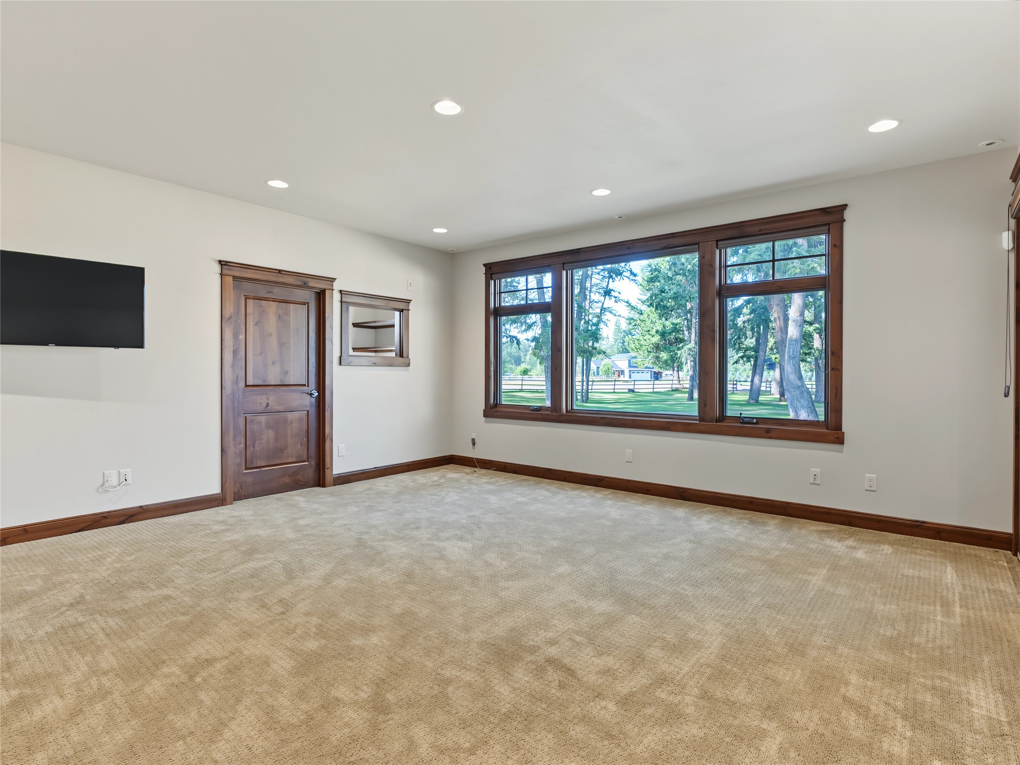 Vacant carpeted room with large wood-framed windows and a wall-mounted television.