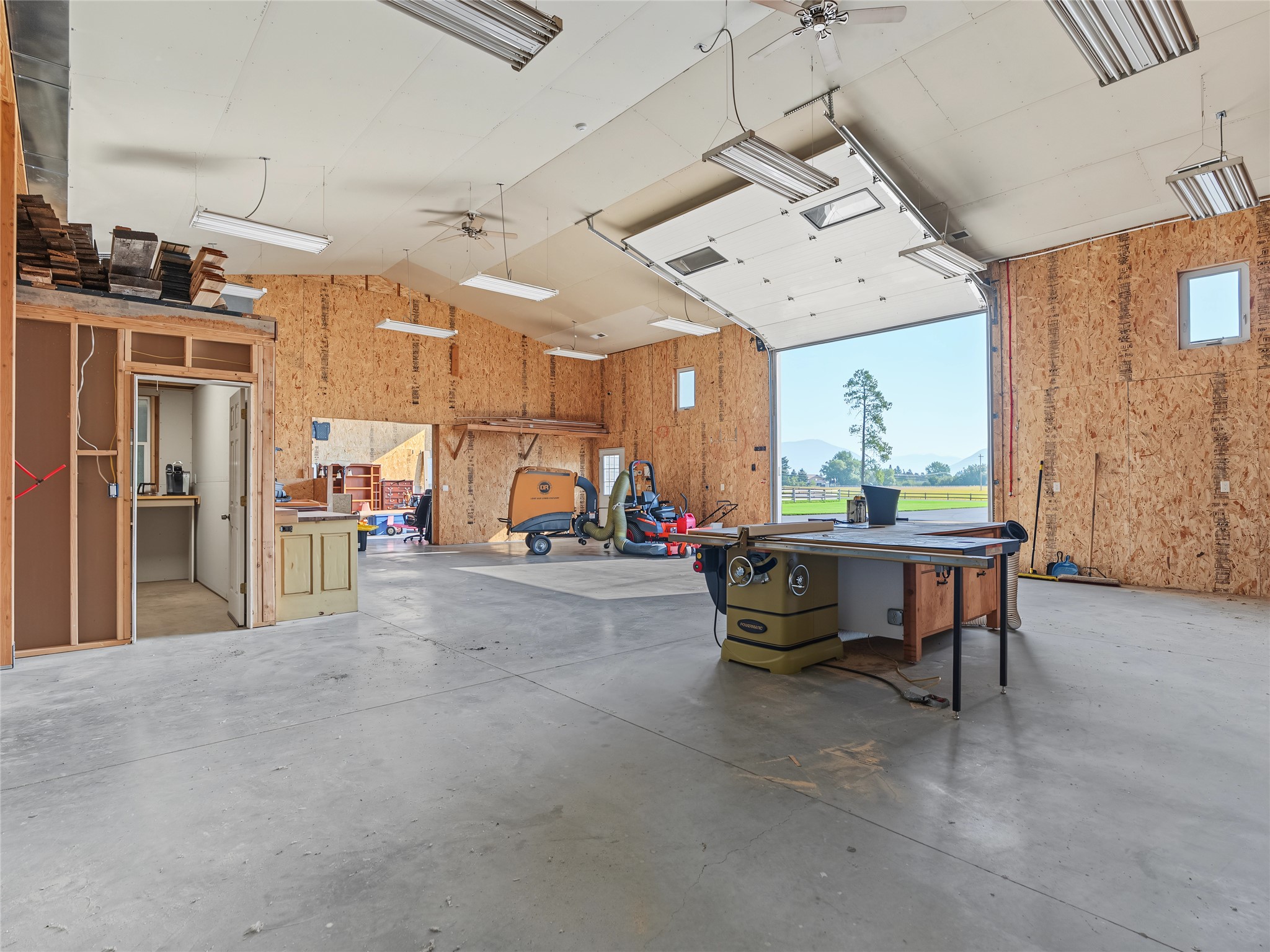 Workshop with woodworking machinery and a large open garage door overlooking a lawn and distant mountains.