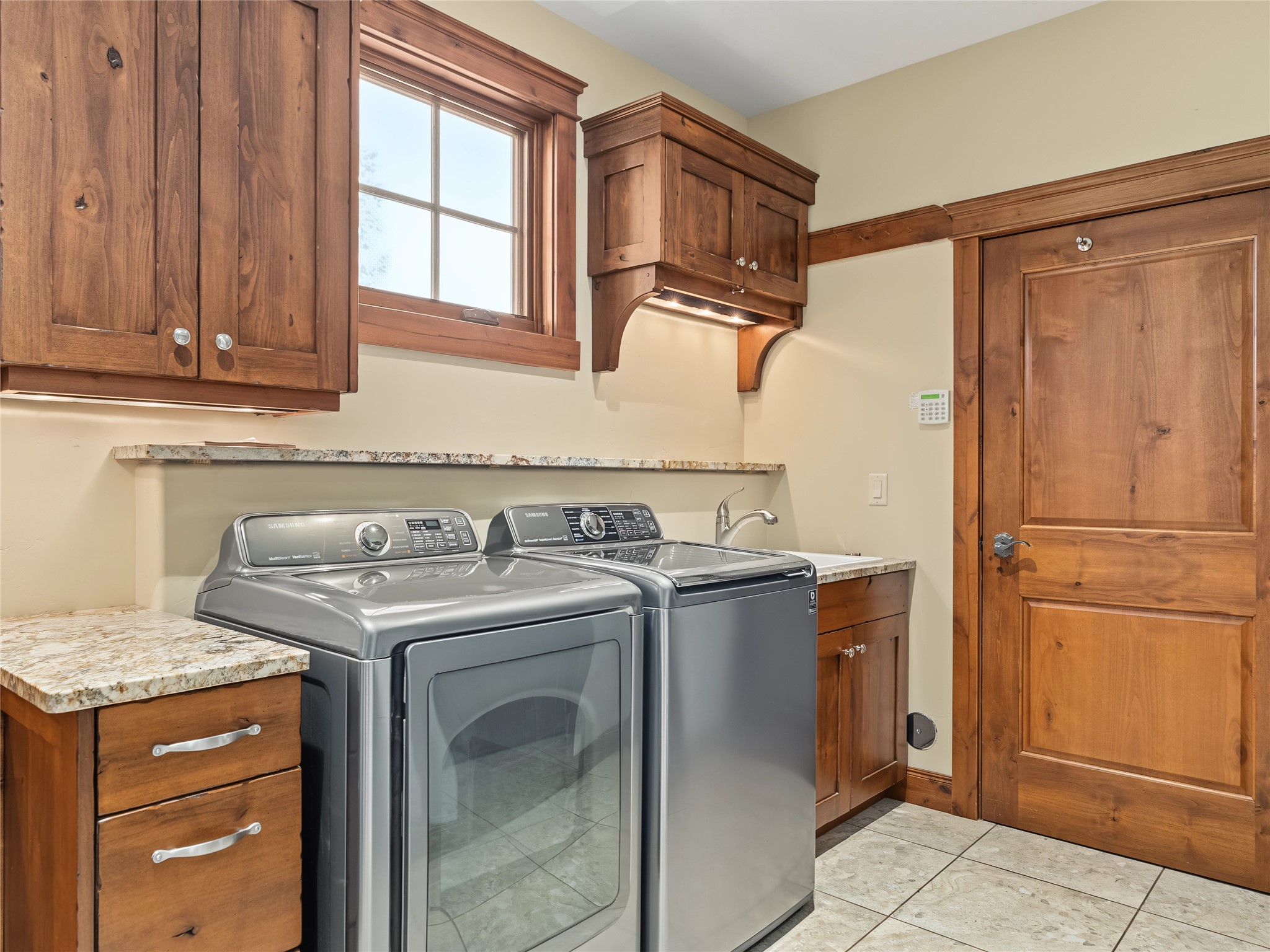 Laundry room with two gray appliances, wood cabinetry, stone counters, and a window.